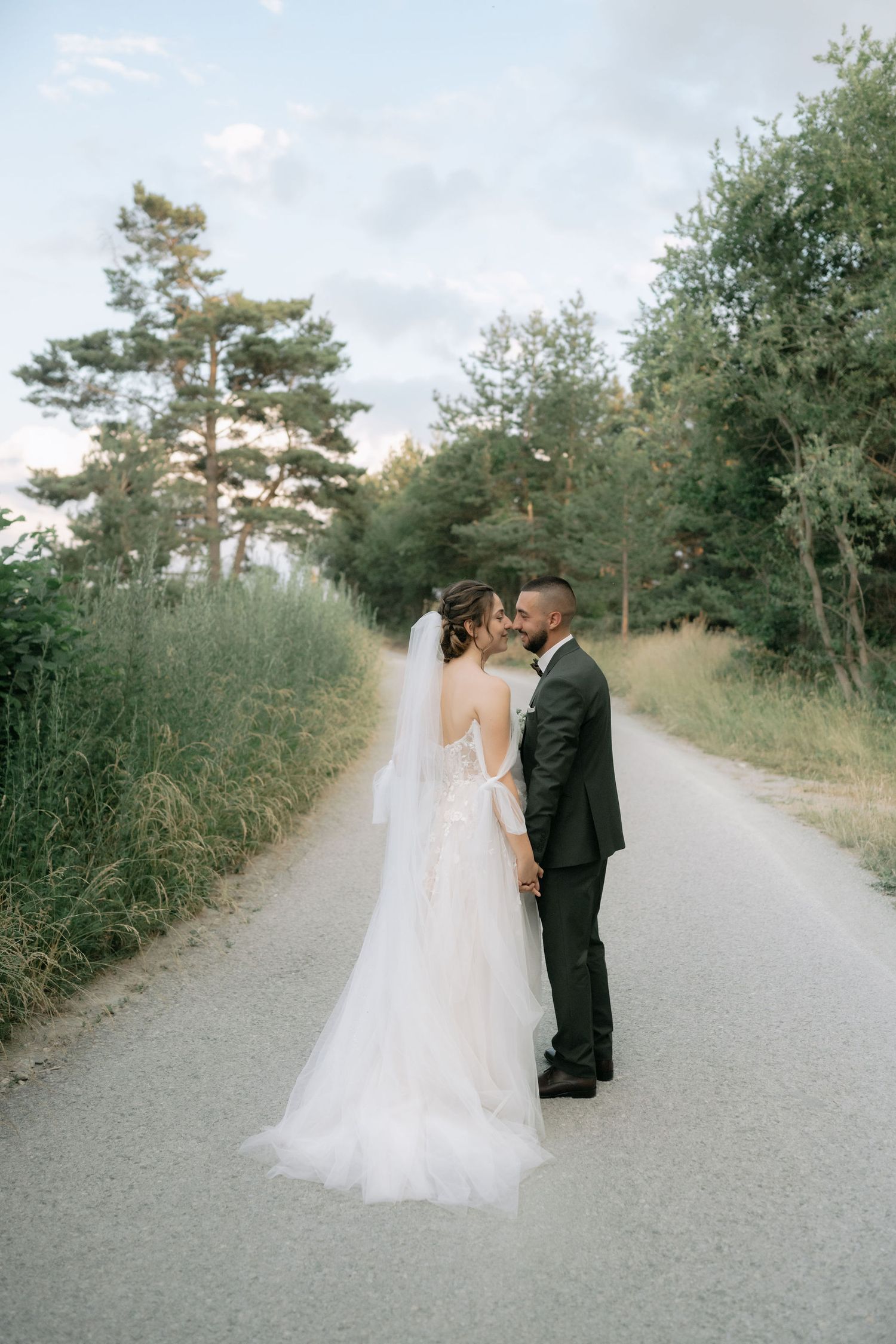 A romantic wedding couple shares intimate moments on a tree-lined gravel path during their outdoor photo session.