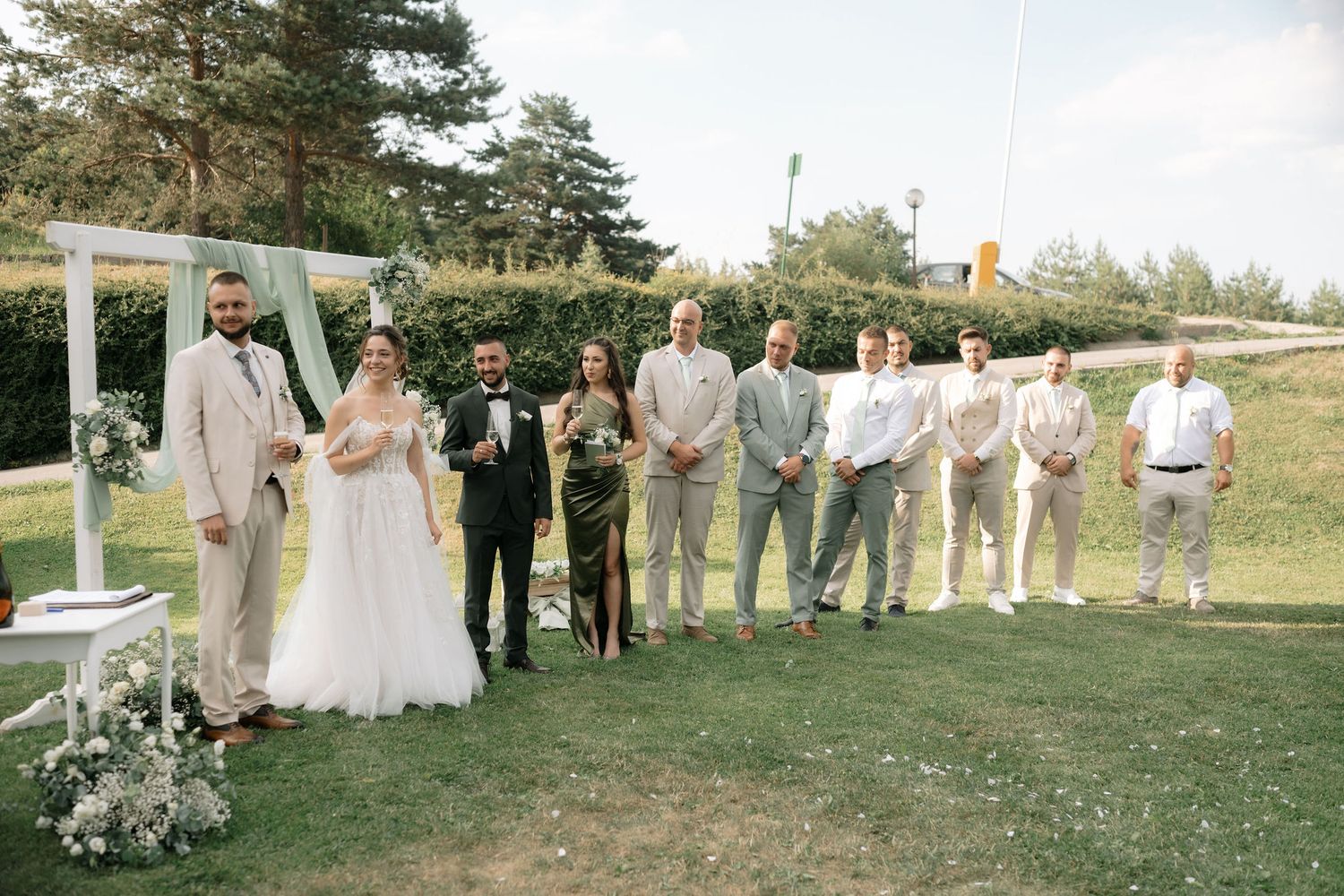 Wedding party members stand in line under white pillars during an outdoor ceremony in a garden setting.