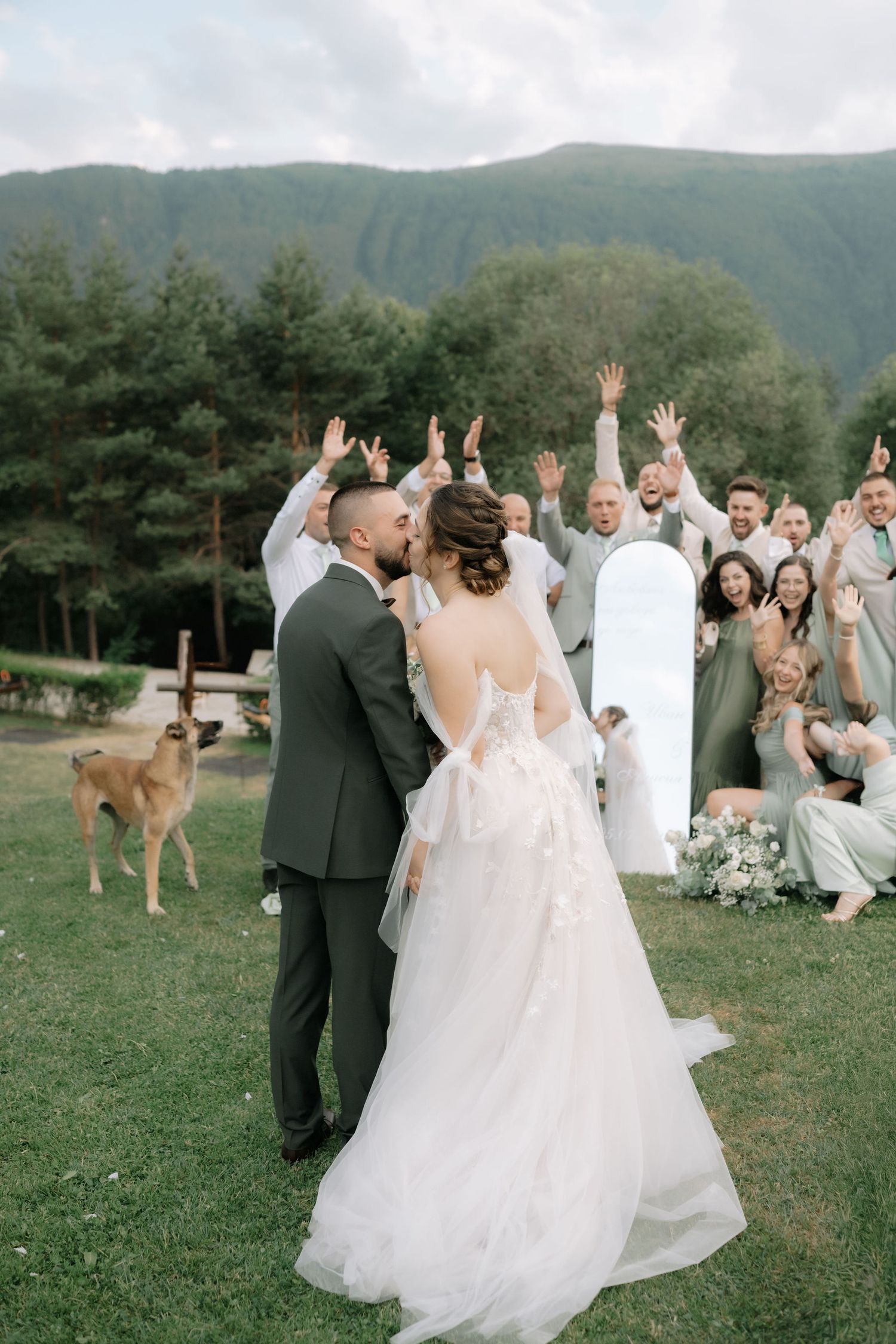 A deer photobombs a joyous outdoor wedding celebration as the wedding party poses for photos in a scenic mountain setting.