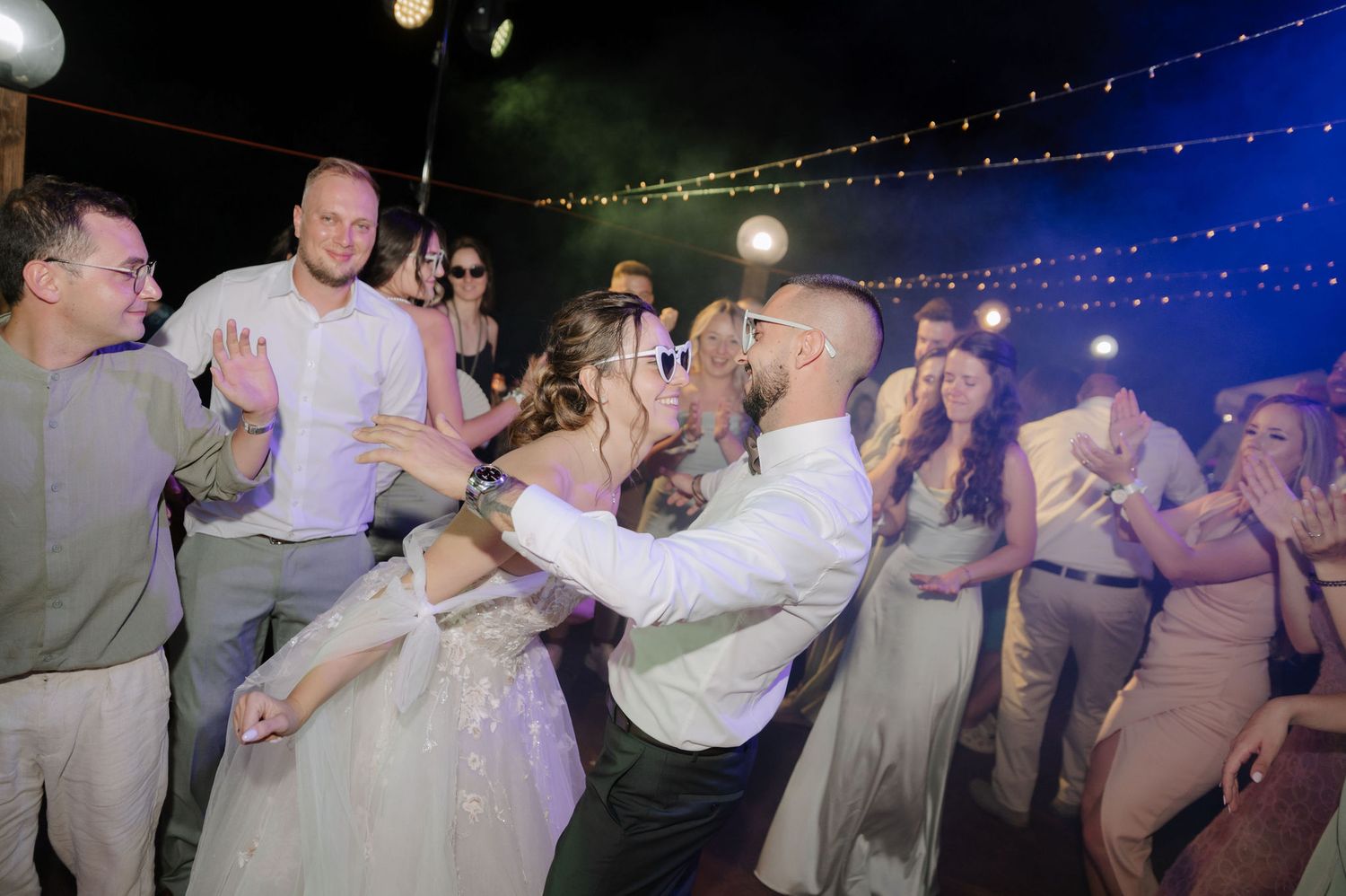 Wedding guests dance together under twinkling lights at an outdoor evening reception celebration.