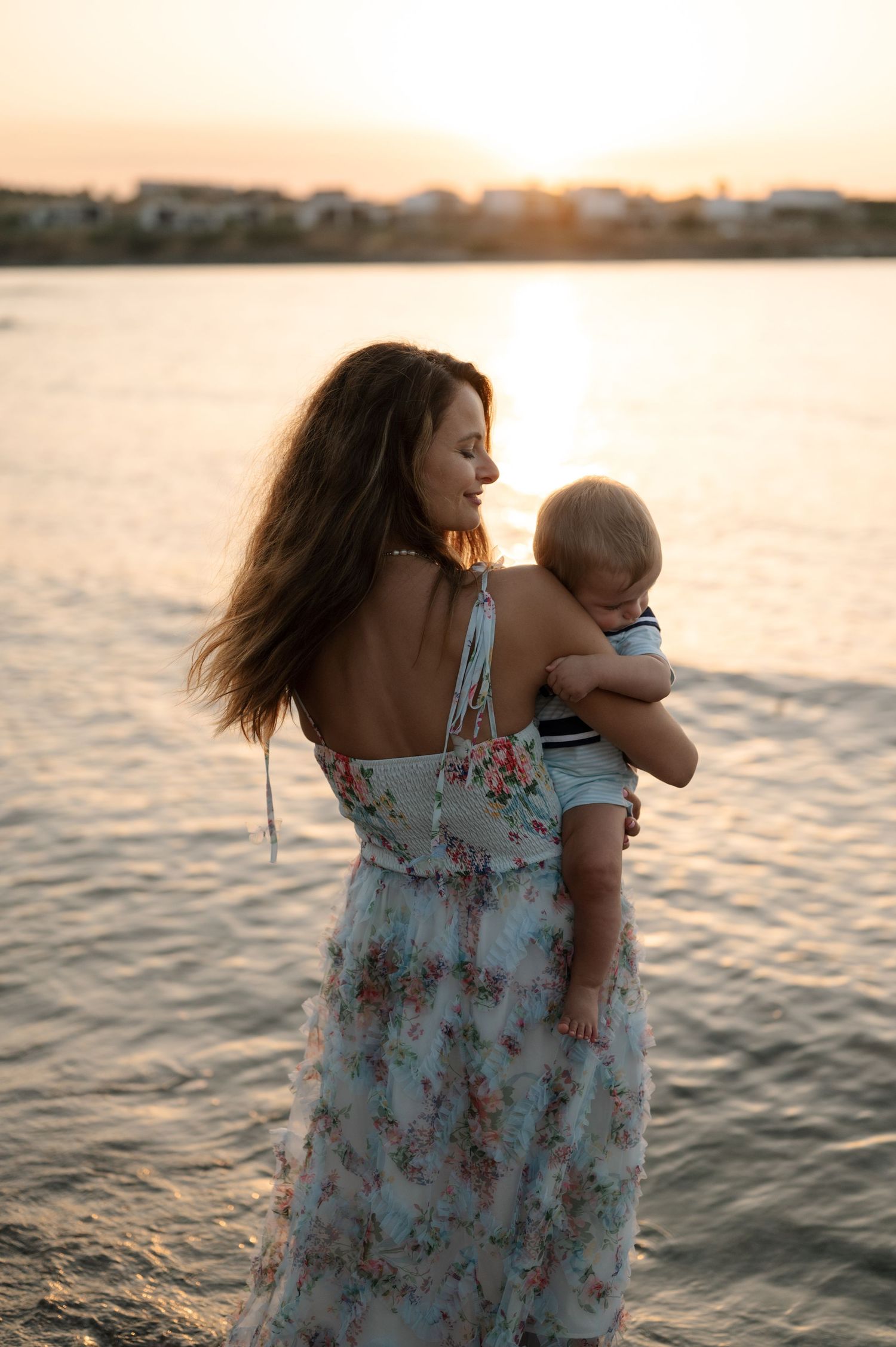 A parent in a floral dress holds a baby while standing by the water at sunset.