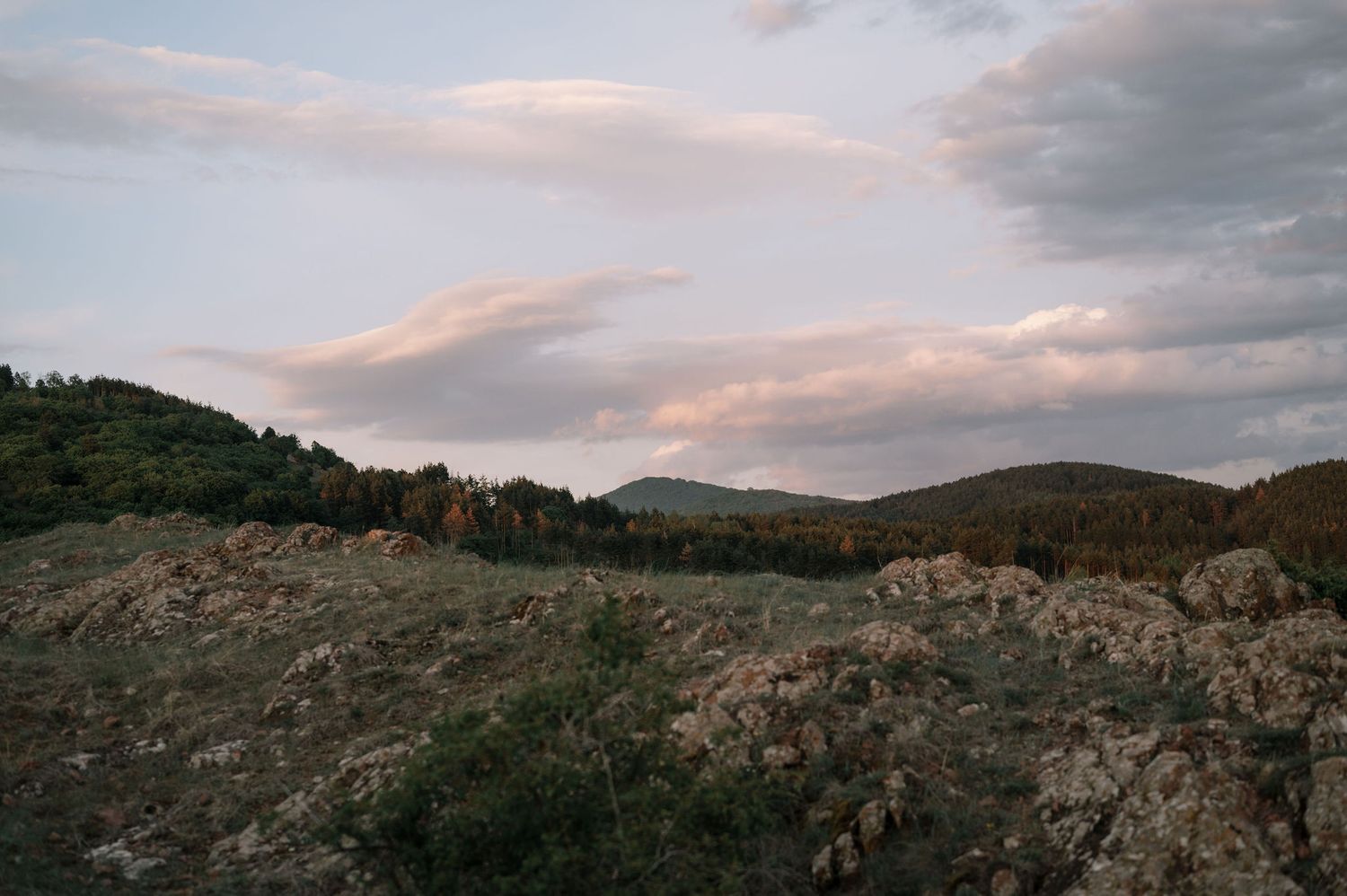A panoramic view of rocky hillsides and mountains under a cloudy sunset sky with warm golden light.