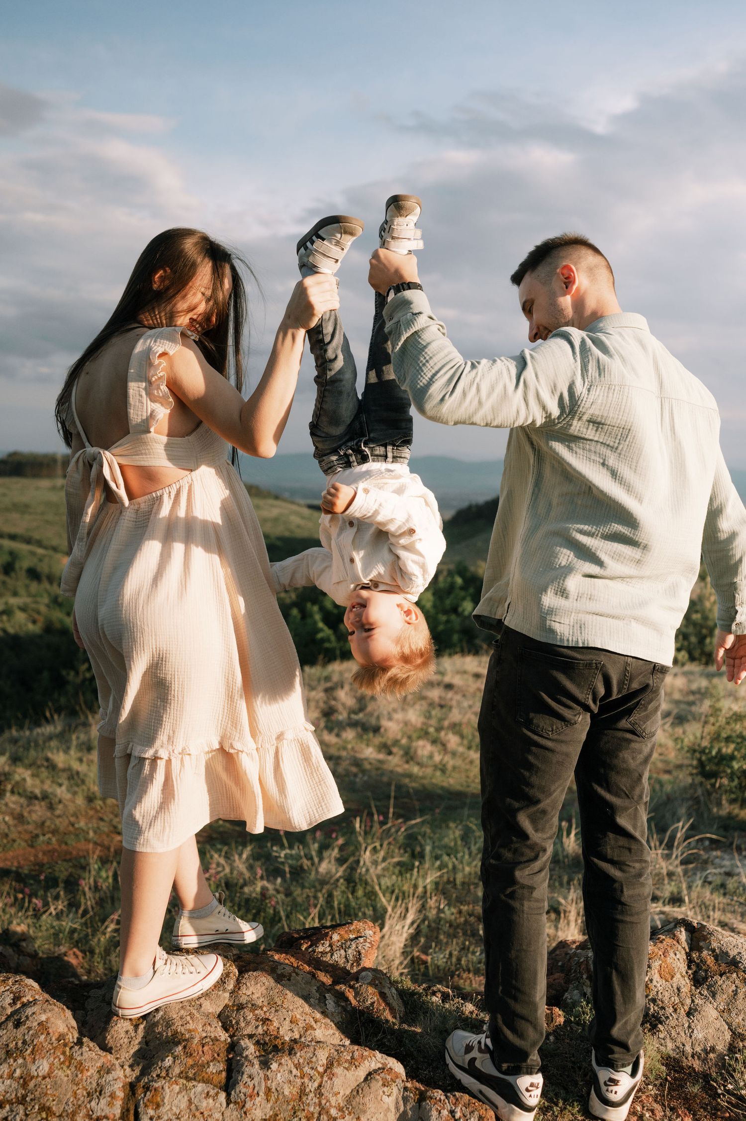 A playful outdoor photoshoot on a hilltop shows a couple lifting their child upside down against a cloudy sky.