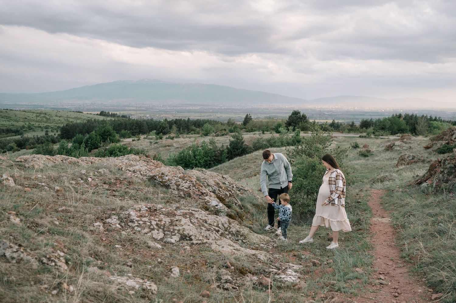 Hikers walk along a winding dirt trail through rugged desert terrain under cloudy skies.