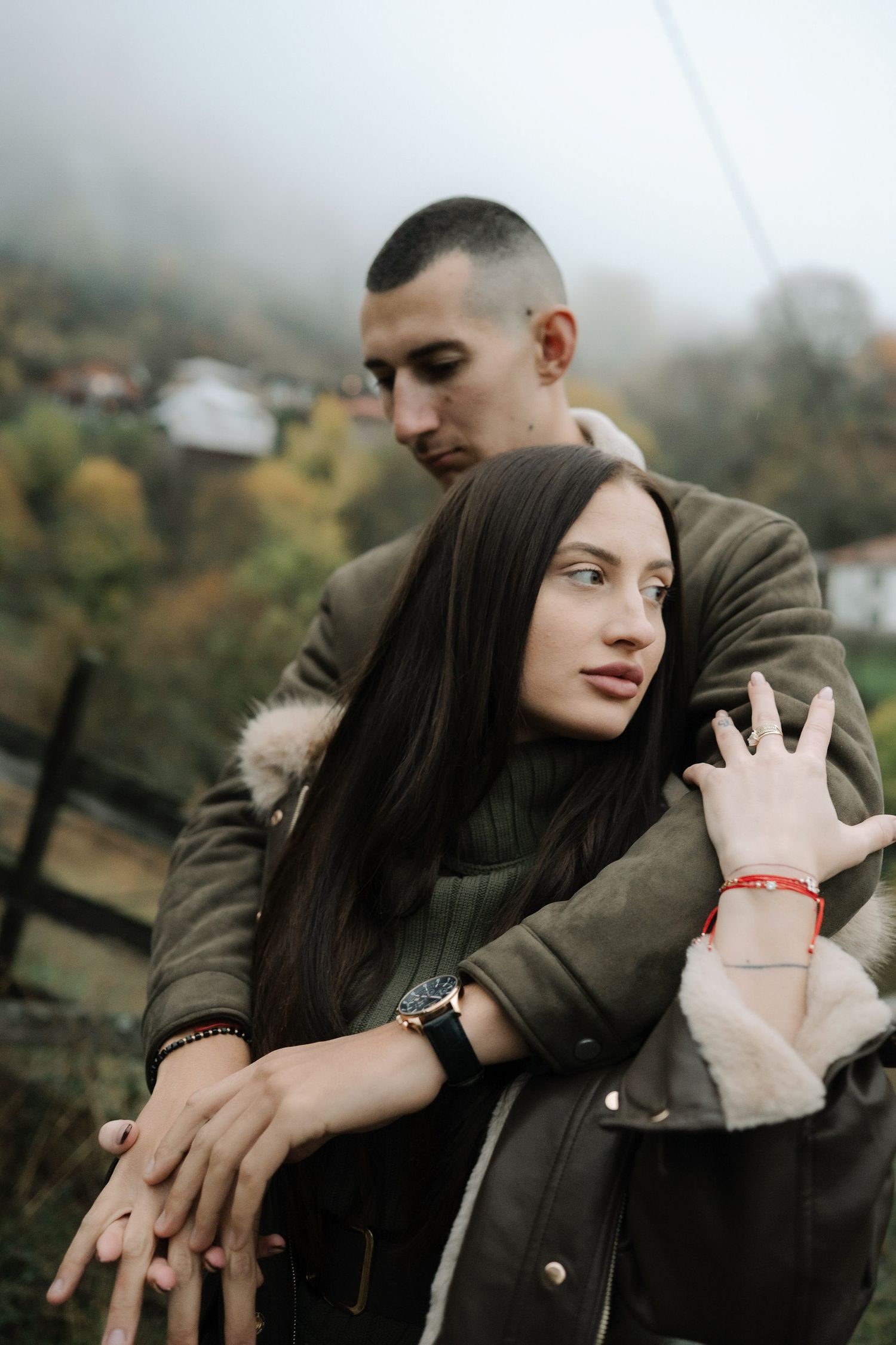 A romantic autumn engagement photo of a couple in matching olive coats against a misty mountain backdrop.
