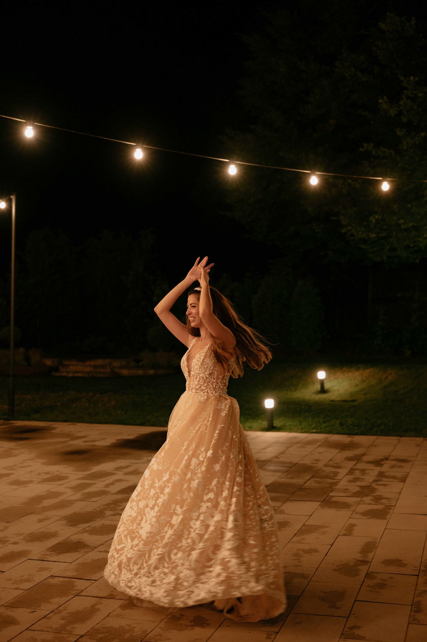 A person in a lace wedding gown twirls on a patio under romantic string lights at night.