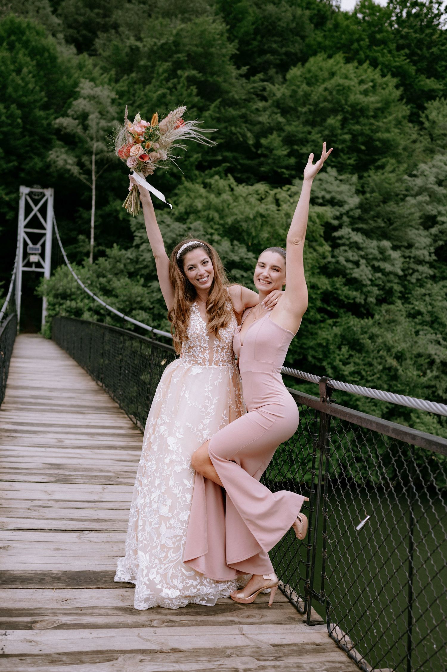 Two friends in pink dresses share joyful moments together on a wooden suspension bridge in a lush forest setting.
