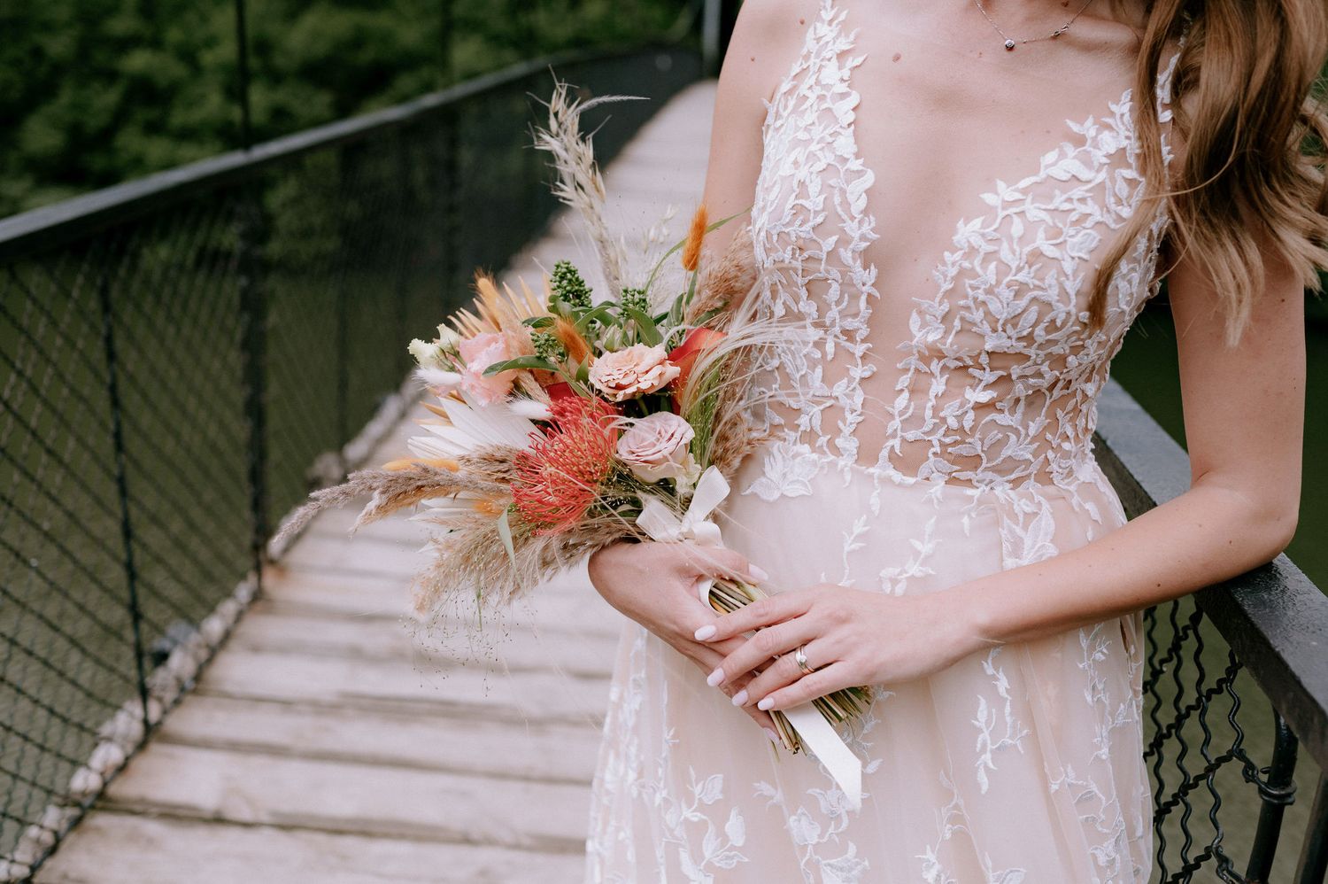 A bride in a delicate lace wedding dress holds a colorful bouquet of flowers on wooden stairs outdoors.