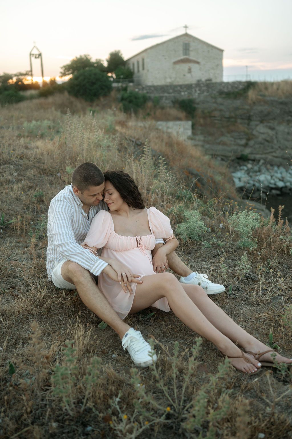 Couple sharing an intimate moment while sitting in a grassy field at sunset with a church visible in the background.