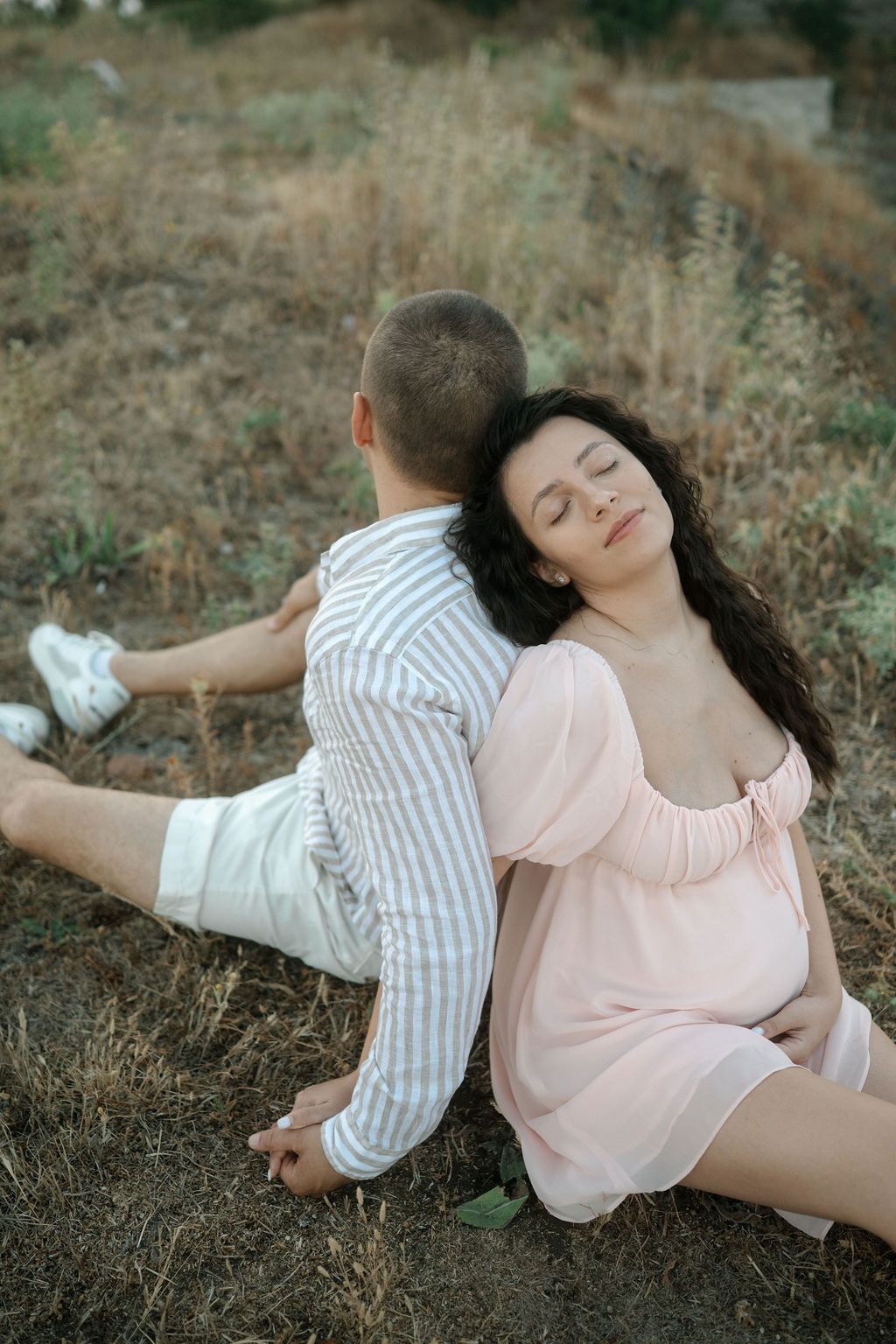 A couple in coordinating pink and striped outfits lay together on dried grass in an artistic photo series.