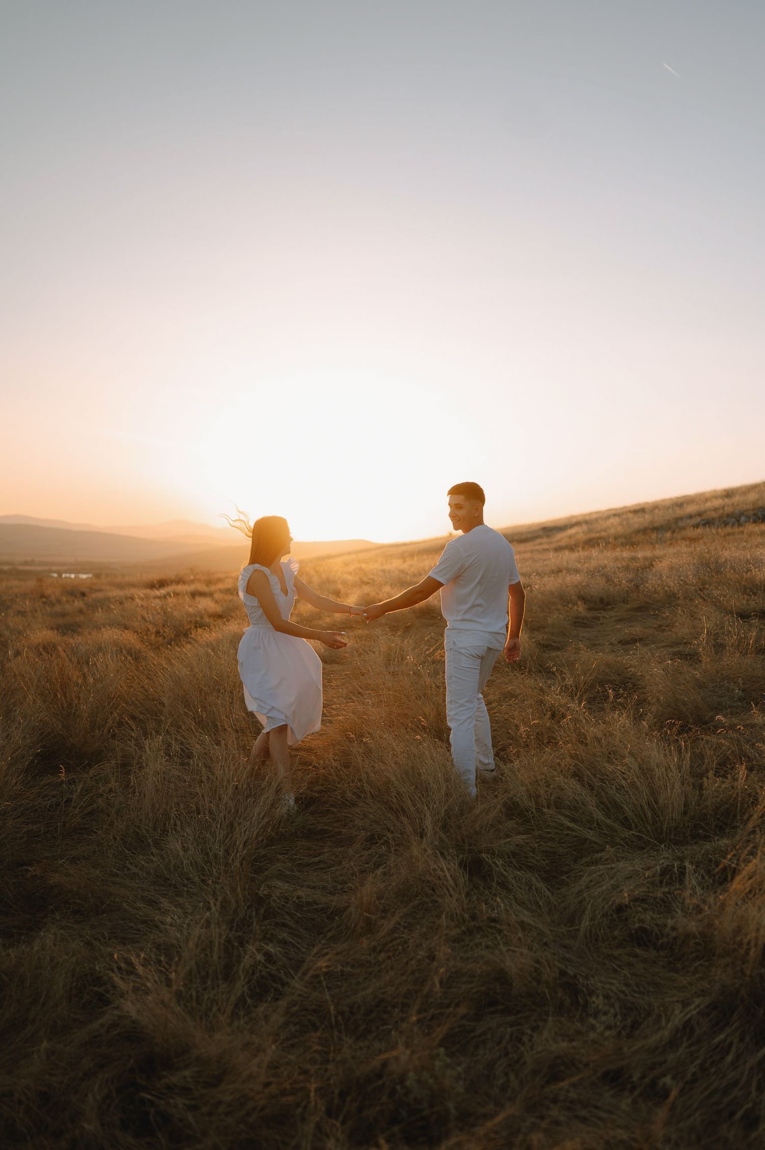 Two silhouettes stand together on a hillside against a warm golden sunset sky.