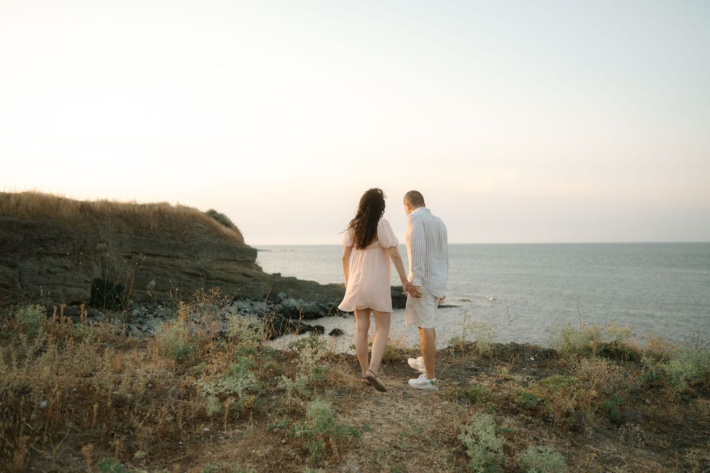 A couple in white outfits walks along a scenic coastal cliff overlooking the ocean at sunset.
