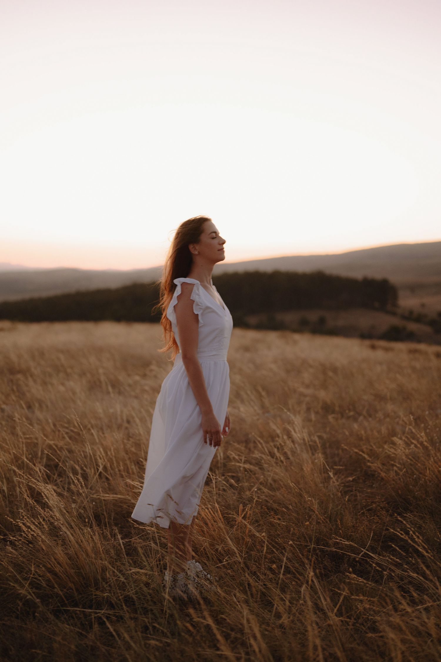A figure in a flowing white dress stands in a golden wheat field at sunset against rolling hills in the background.