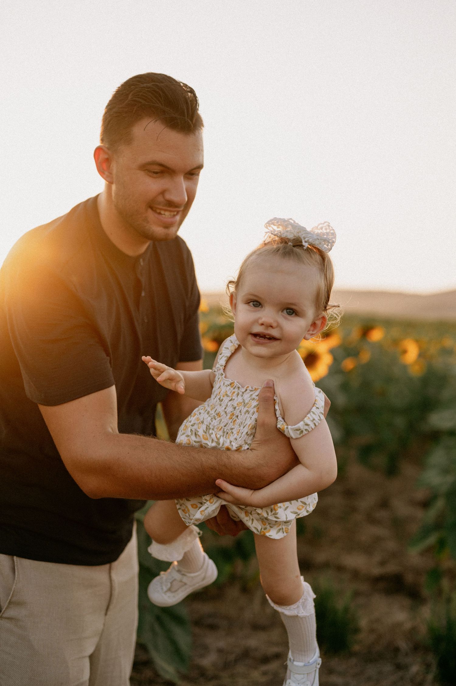 A smiling man holds a baby girl in a floral romper and bow headband in a sunflower field at golden hour.