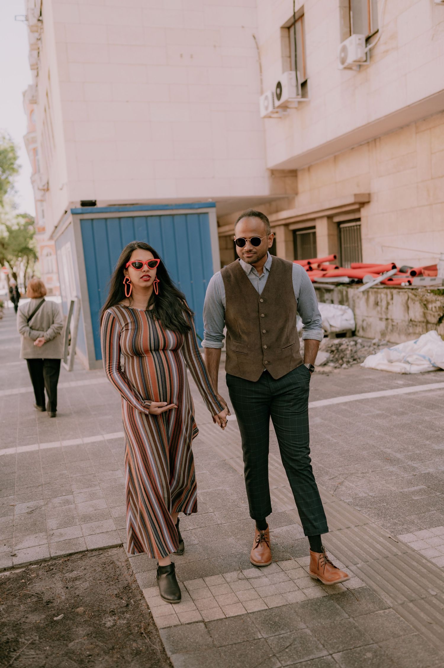 Stylish couple walking hand-in-hand on a city sidewalk, woman in striped maxi dress, man in vest and trousers.