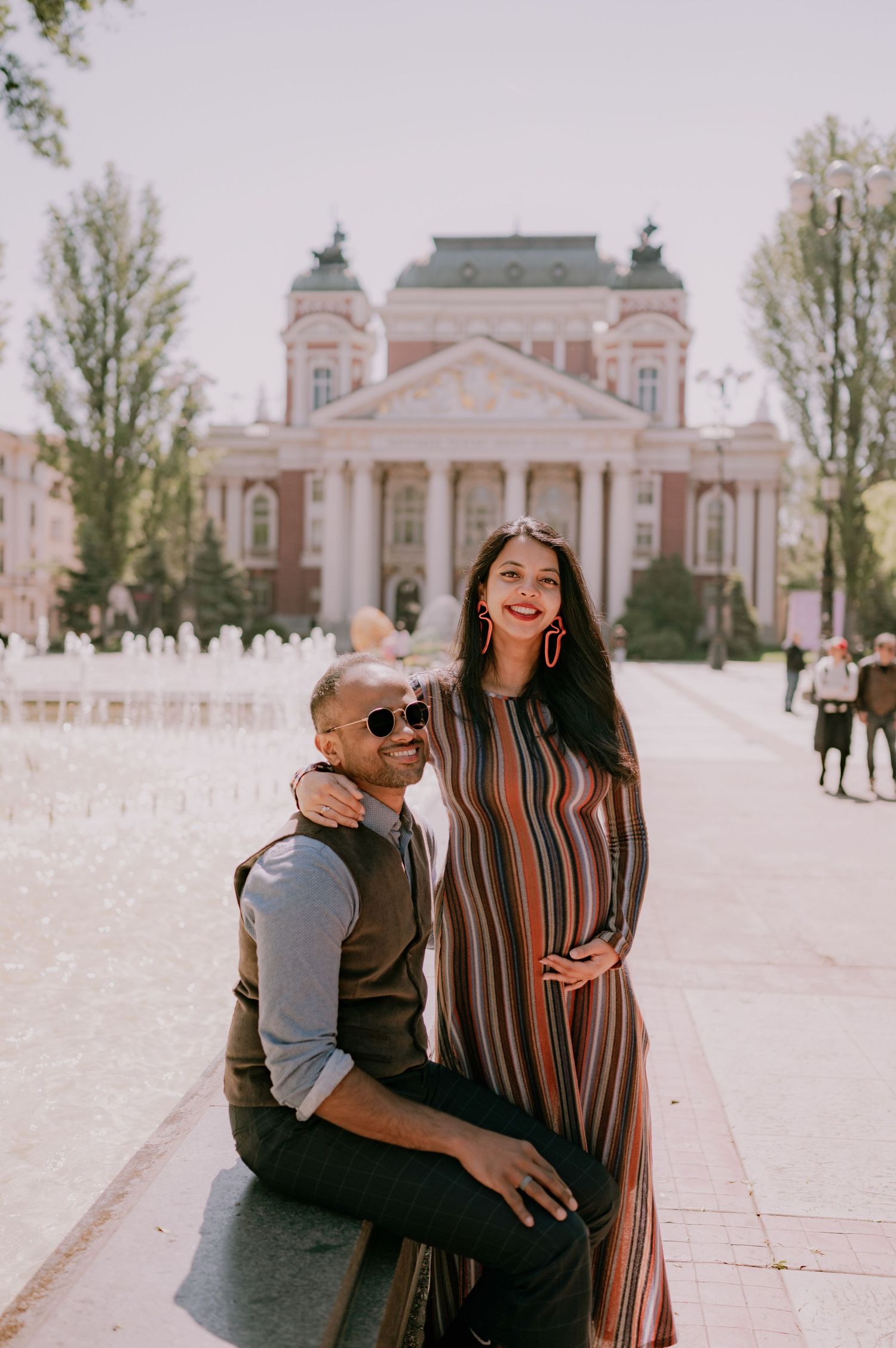 Couple posing in front of a grand neoclassical building, woman in striped dress touching her baby bump, sunny day.