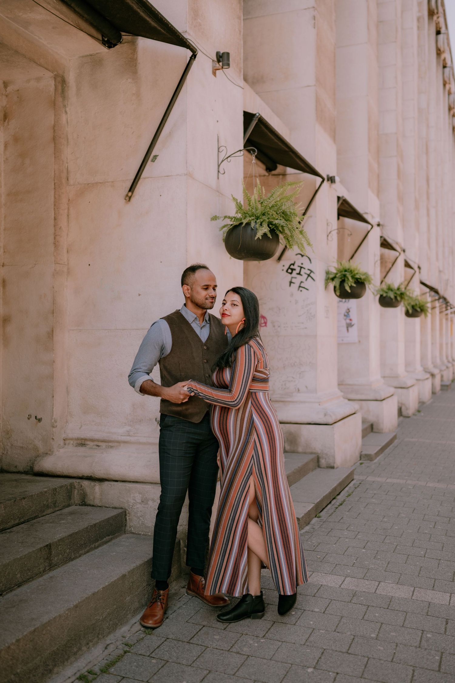A couple embraces in front of a grand stone building with hanging fern planters on a cobblestone street.