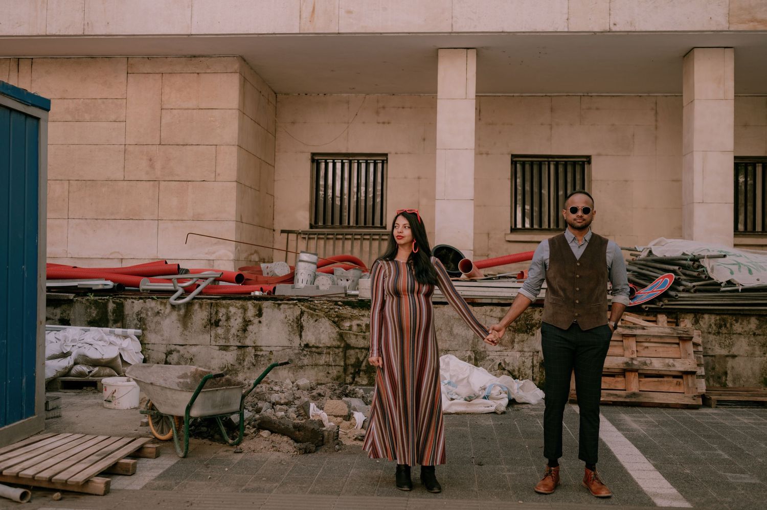 Couple holding hands in front of a rustic stone wall with scattered materials in an urban outdoor setting.