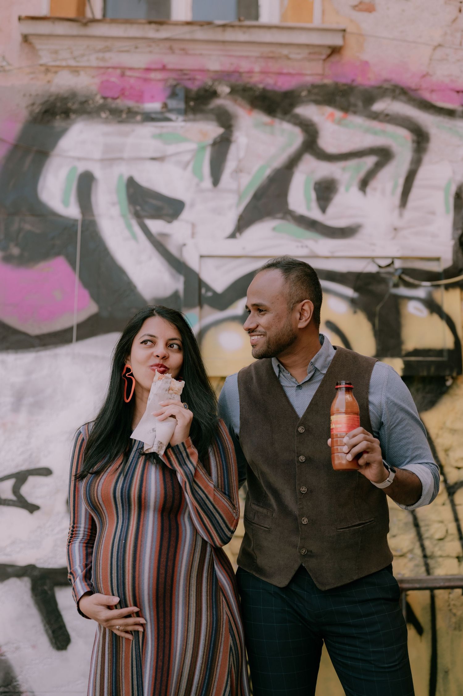 Pregnant woman in striped dress and man in vest pose before graffiti wall, she holds food, he holds a juice bottle.