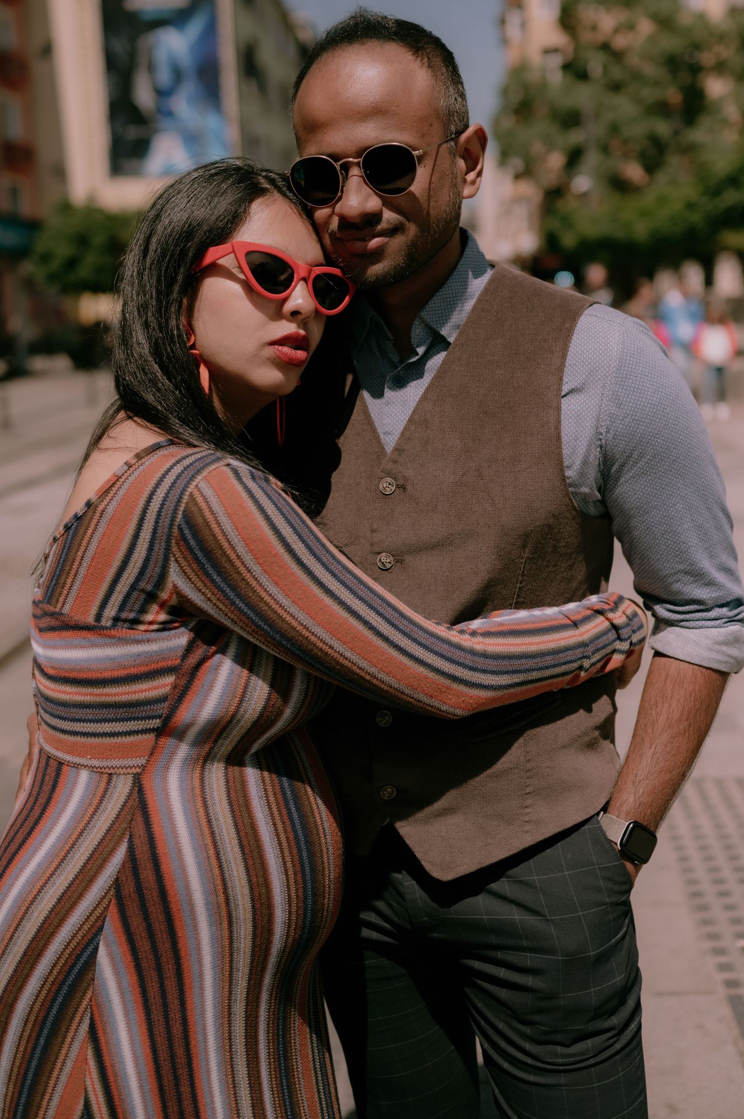 A stylish couple embracing on a city street, woman in striped dress with red sunglasses, man in vest and round shades.