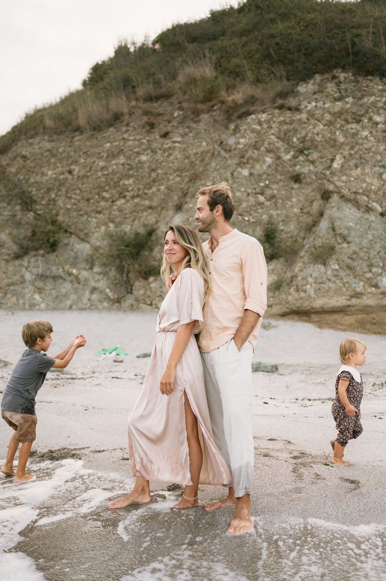 A couple stands barefoot on a sandy beach while two young children play nearby with rocky cliffs in the background.