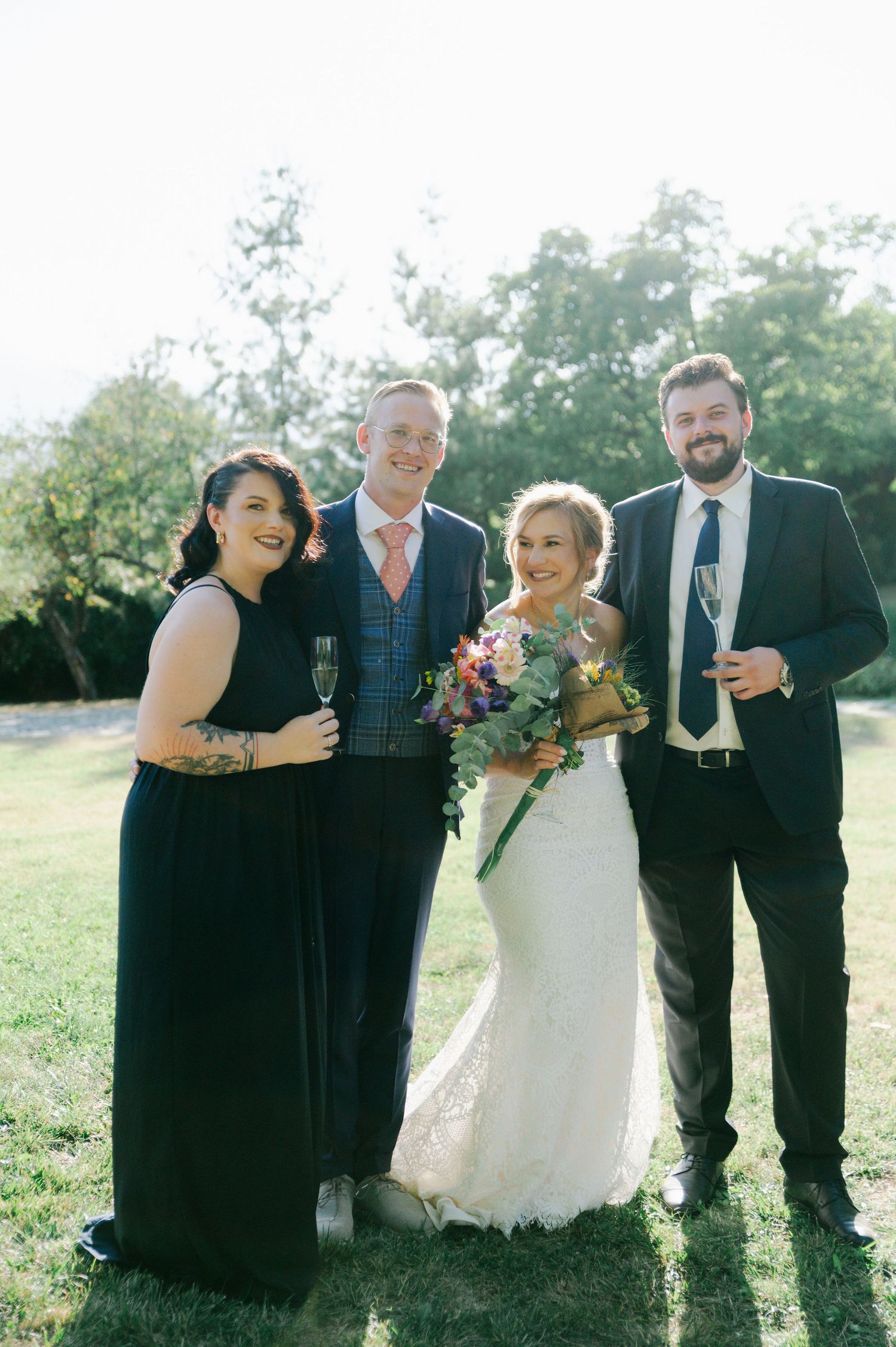Wedding group shares laughs while posing outdoors in formal black attire and white dress.