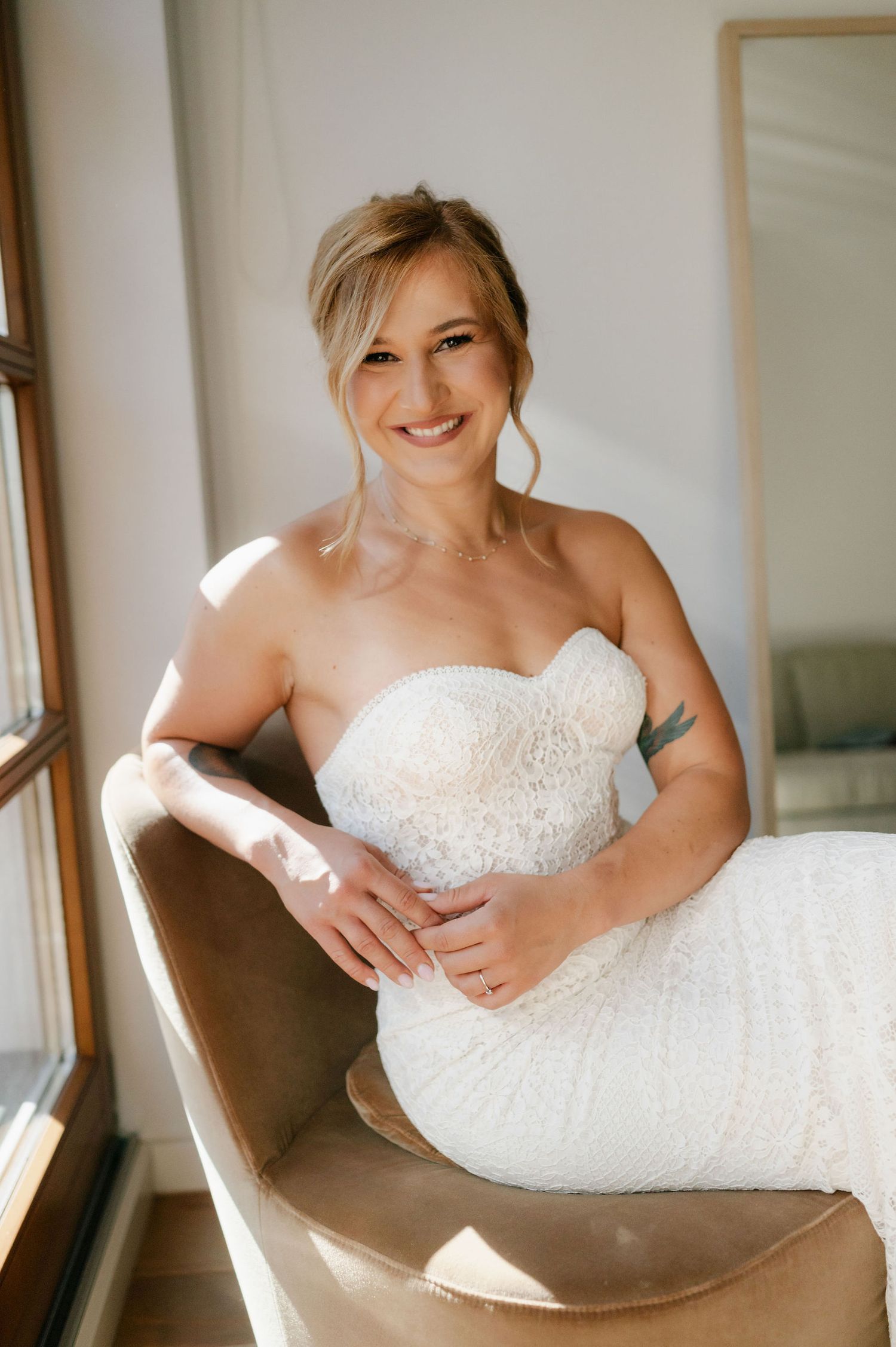 A wedding dress creates elegant curves while posed in a modern brown chair by a window.