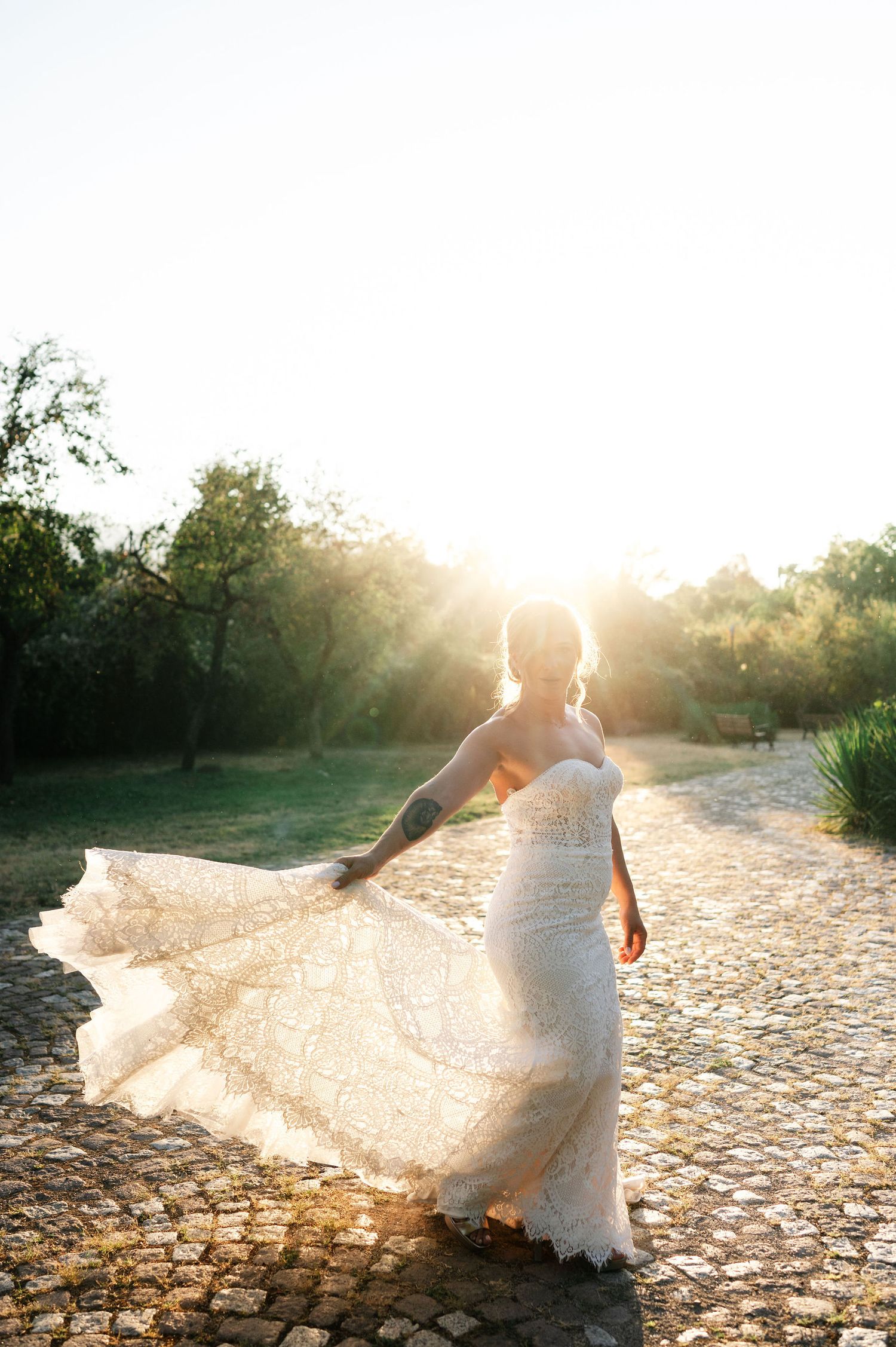 Bride twirls in lace wedding dress on cobblestone path during sunset.