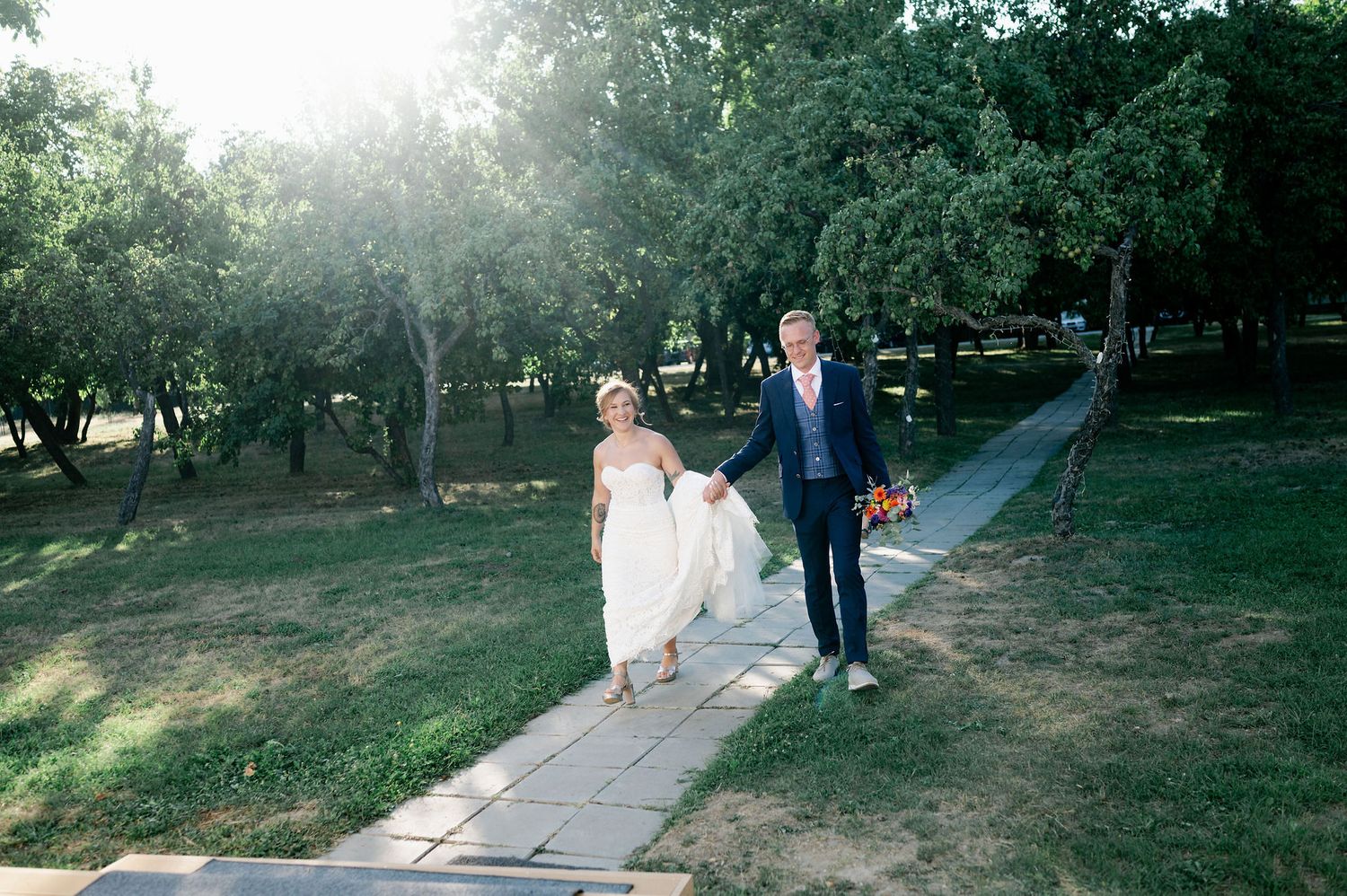 Wedding couple walking together on a stone path through a park with golden sunlight filtering through trees.