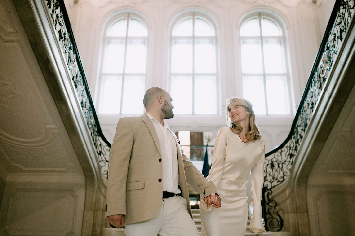 A couple in cream-colored outfits share a romantic moment on an elegant staircase with ornate railings.