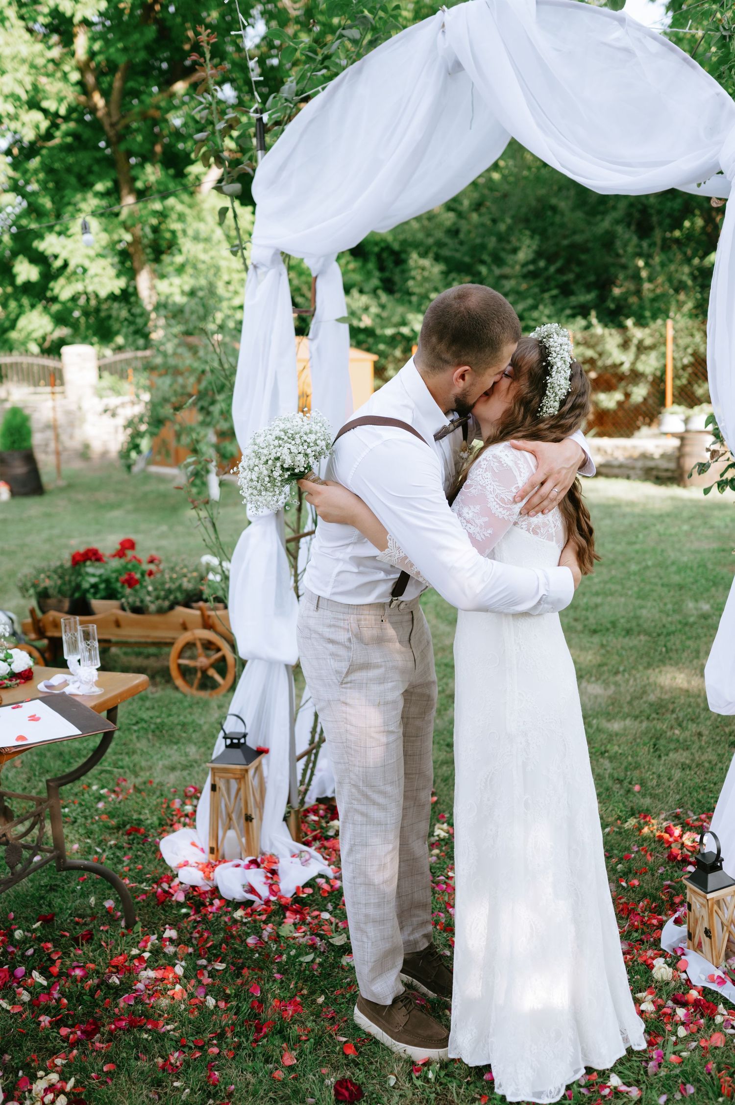 A romantic outdoor wedding ceremony under a white draped arch with floral decorations and rustic decor in a garden setting.