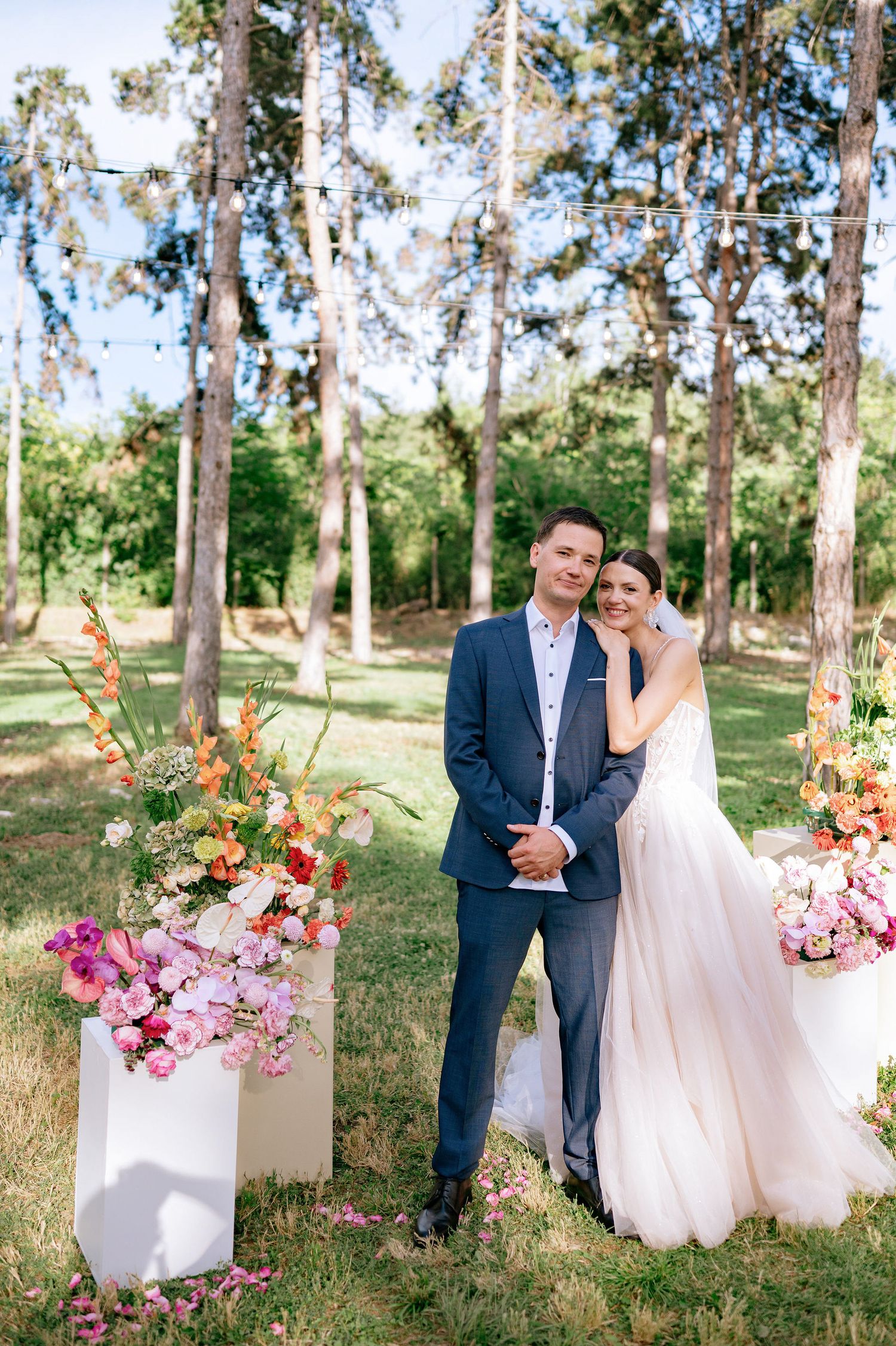 A romantic outdoor wedding ceremony setup in a wooded area with tall trees and floral arrangements in white stands.