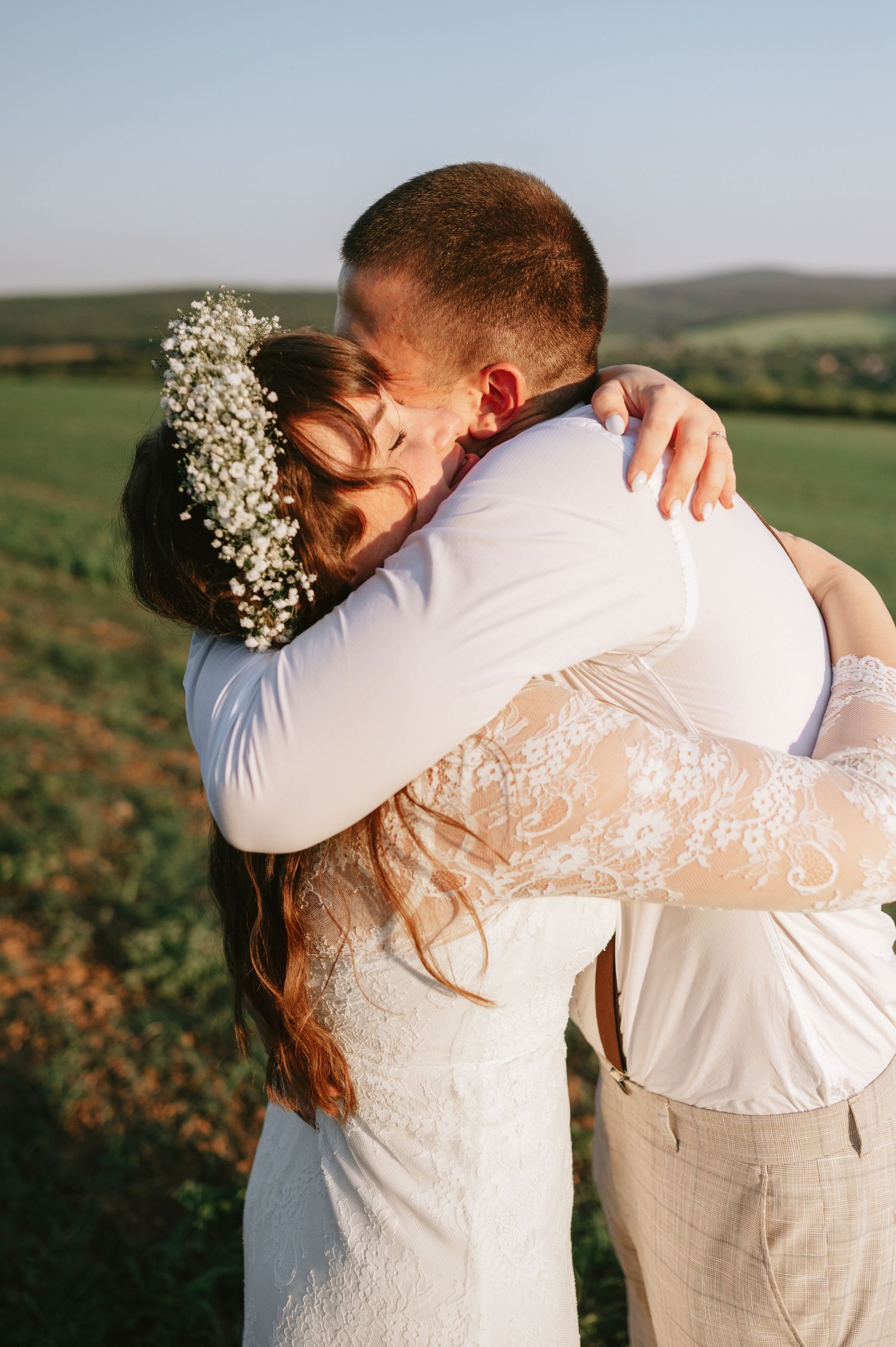 An intimate embrace between newlyweds in white wedding attire against a natural outdoor backdrop.