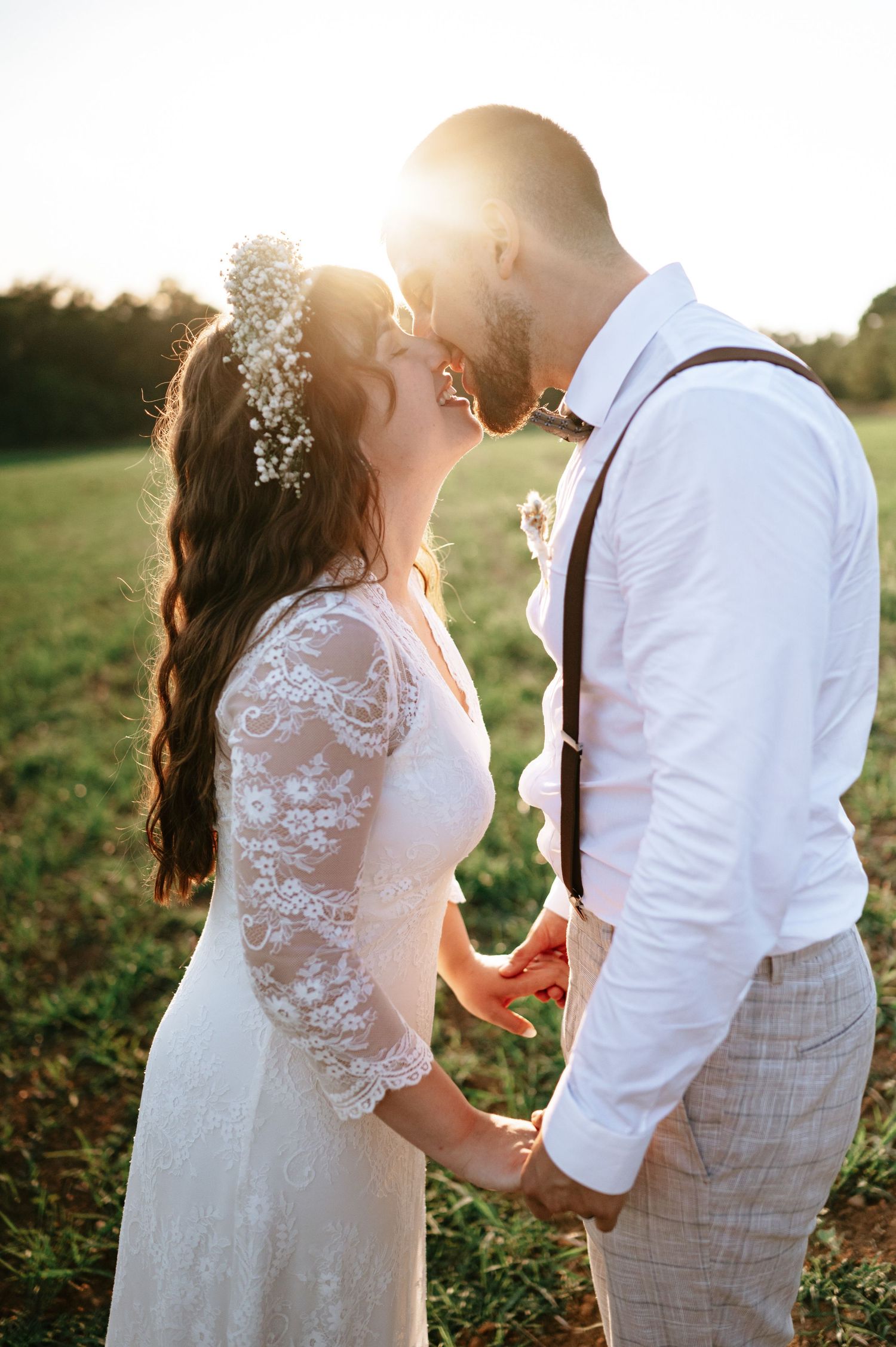 Bride in lace dress and groom in suspenders hold hands during sunset wedding photos.