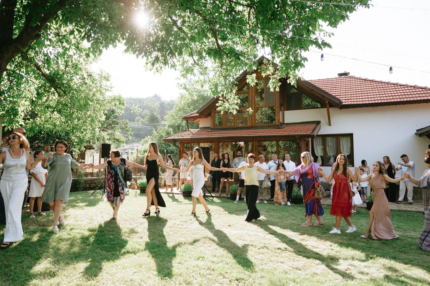 Wedding guests gathered outside a rustic venue under tall trees in late afternoon light.