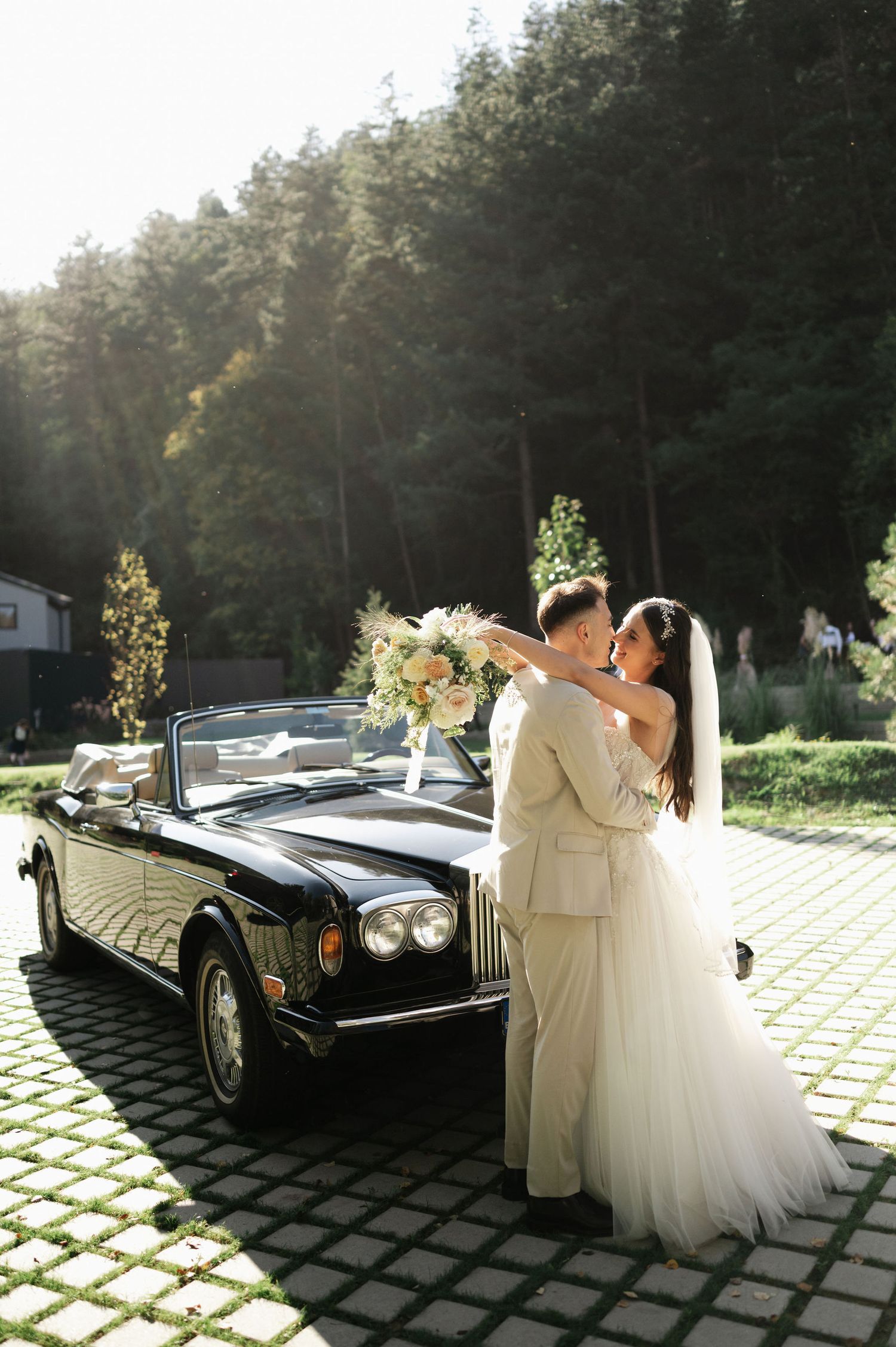 A romantic wedding moment captured next to a vintage black car on a sunny afternoon.