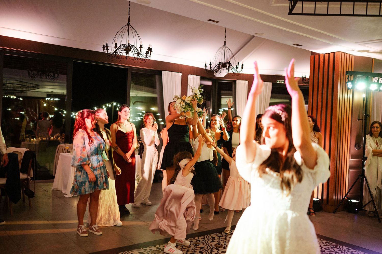 A group of wedding guests dance and celebrate under chandeliers in a reception venue.