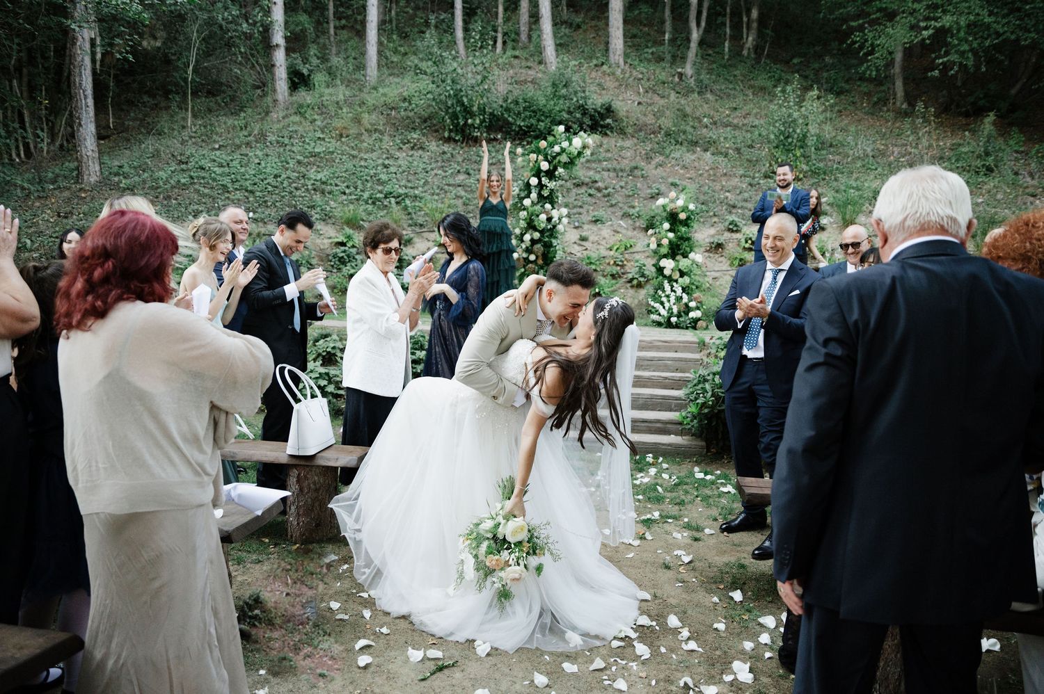 Outdoor wedding ceremony in progress with guests gathered among trees and stone steps.