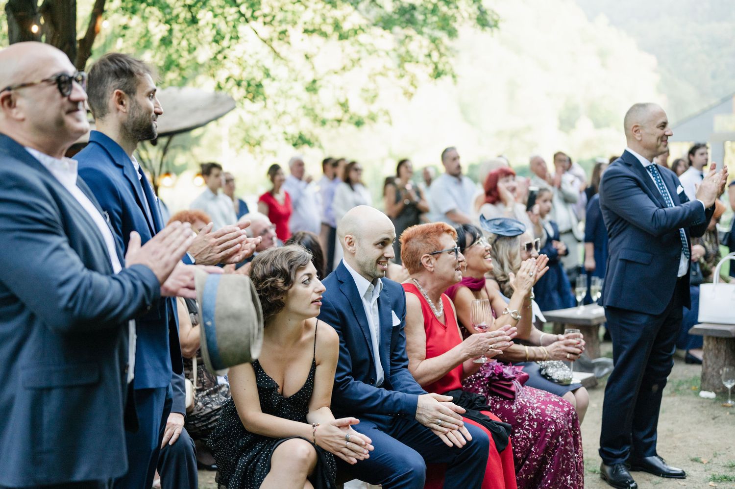 Wedding guests seated outdoors clap during ceremony in forest setting.