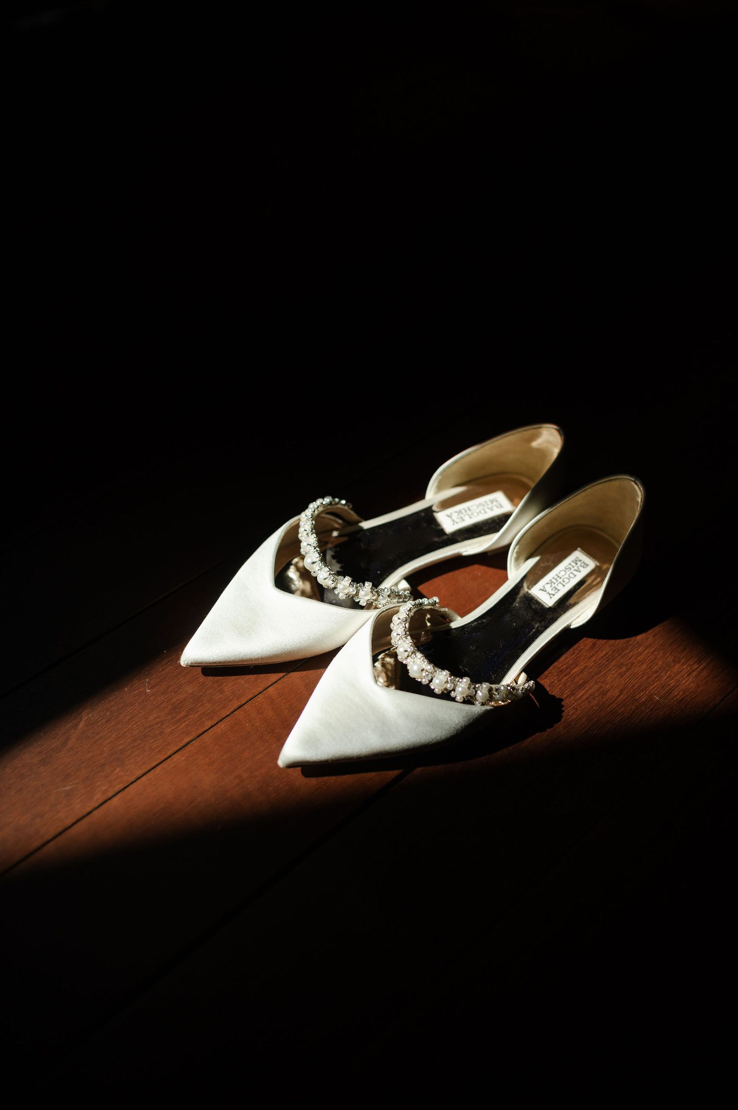 White bridal shoes with pearl details photographed dramatically on dark wooden surface.
