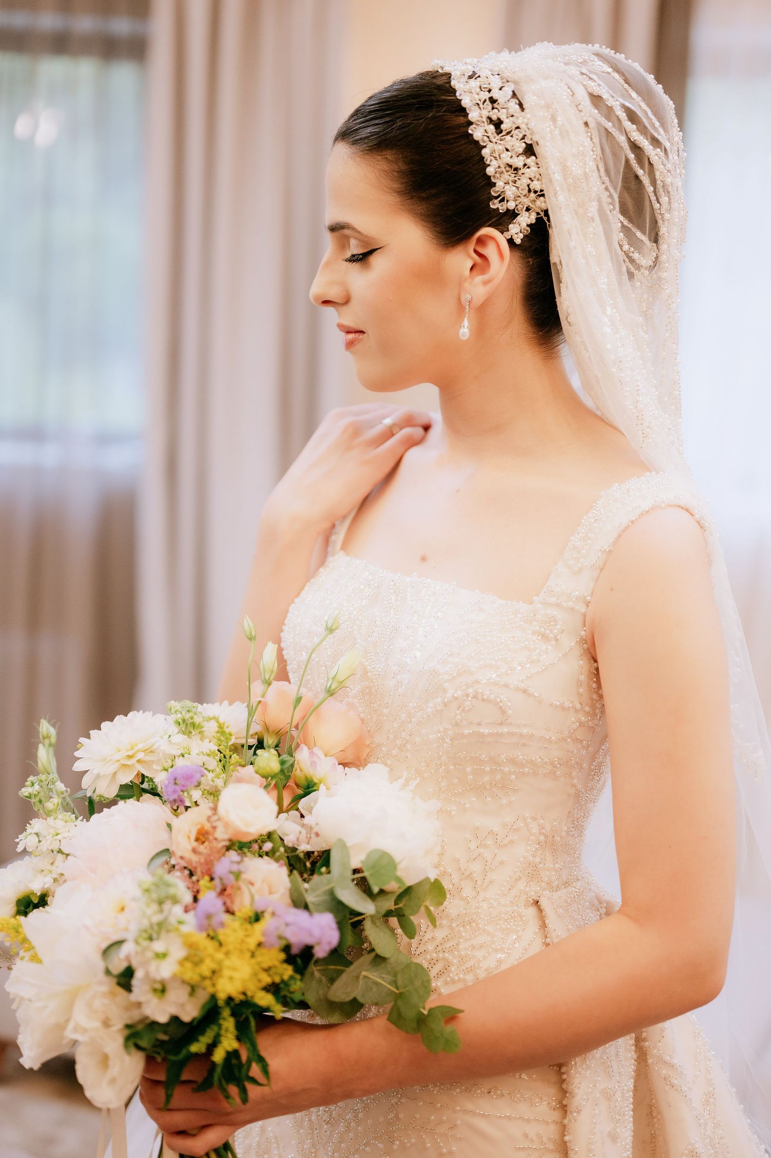 Elegant bride in lace dress with pearl headpiece holding romantic pastel floral bouquet.