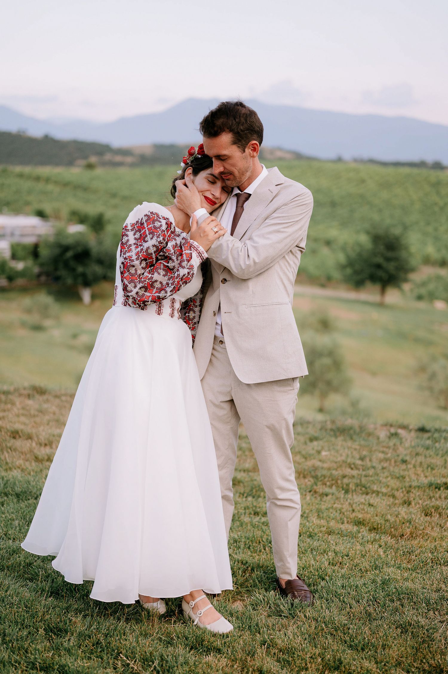 Wedding couple shares emotional moment on hillside wearing traditional dress with red embroidered details.