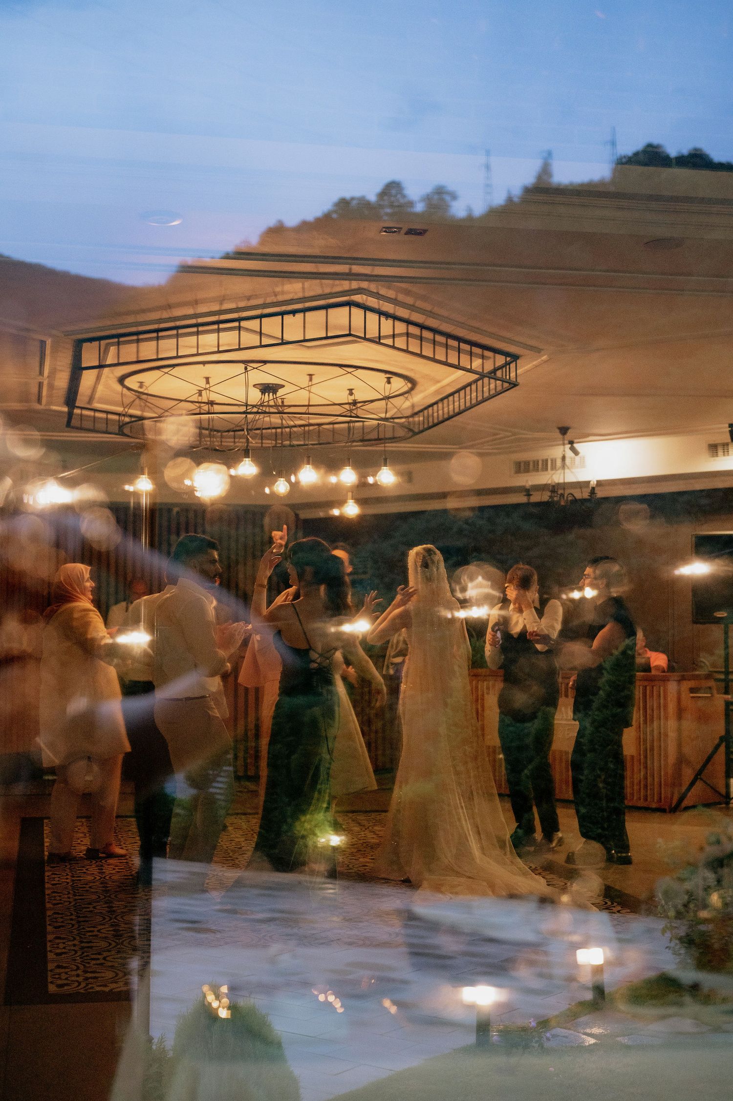 Motion-blurred scene of wedding guests dancing under string lights at dusk.