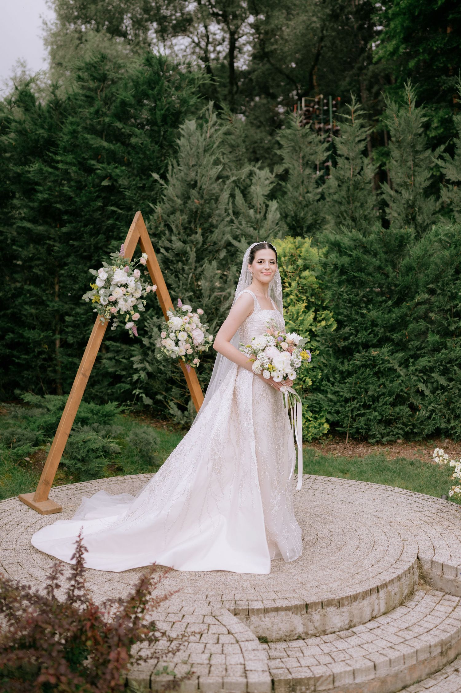 Bridal portrait on a circular stone patio surrounded by evergreen trees and wooden ceremony arch.