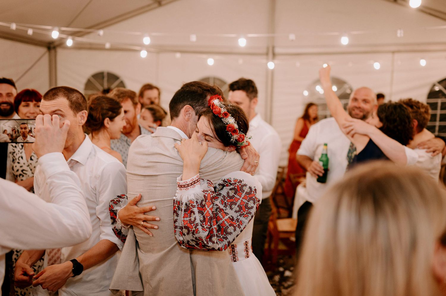 Wedding guests dancing and celebrating under warm string lights in reception tent.