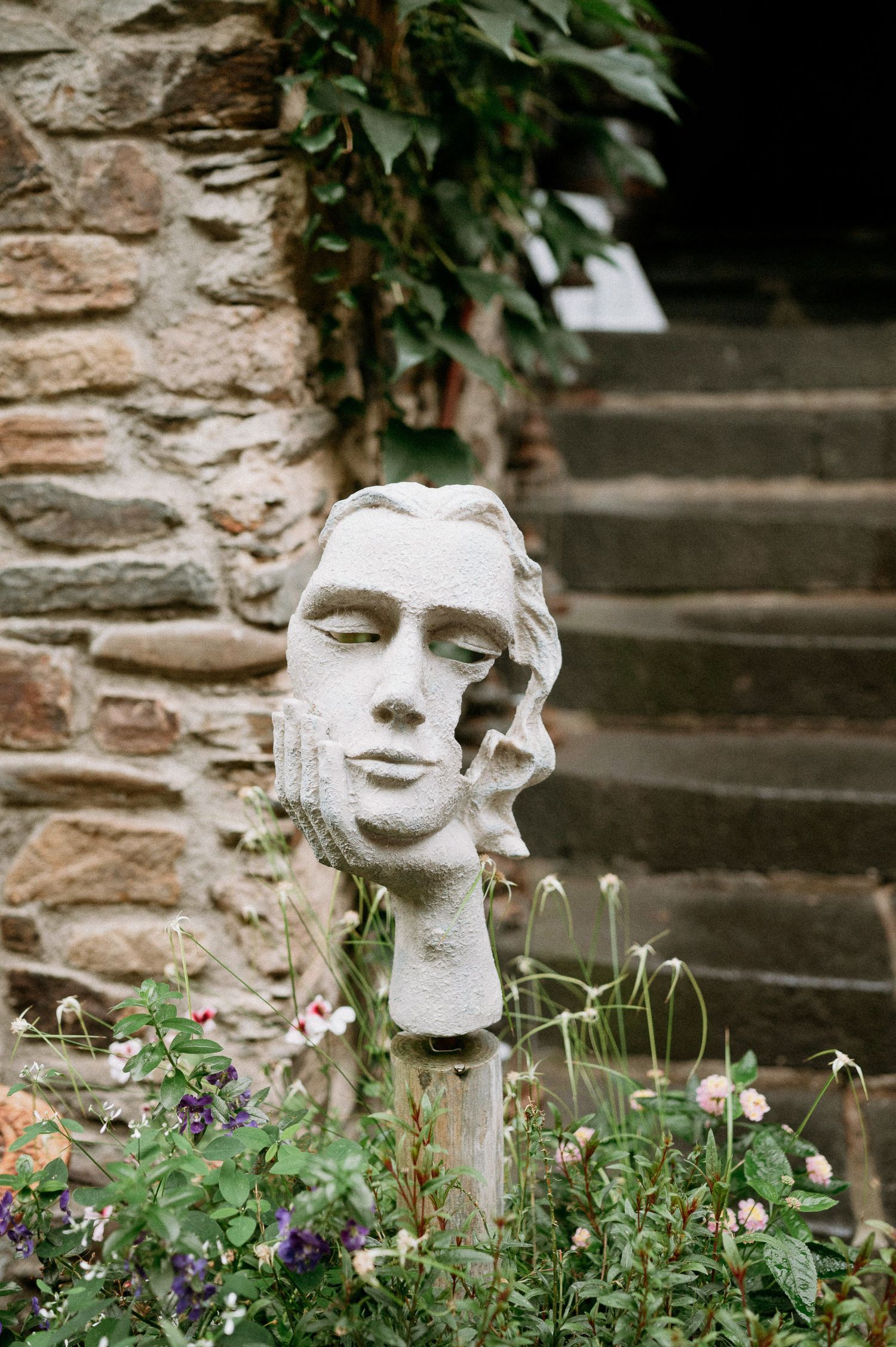 A classical stone sculpture head among garden flowers and stone steps.