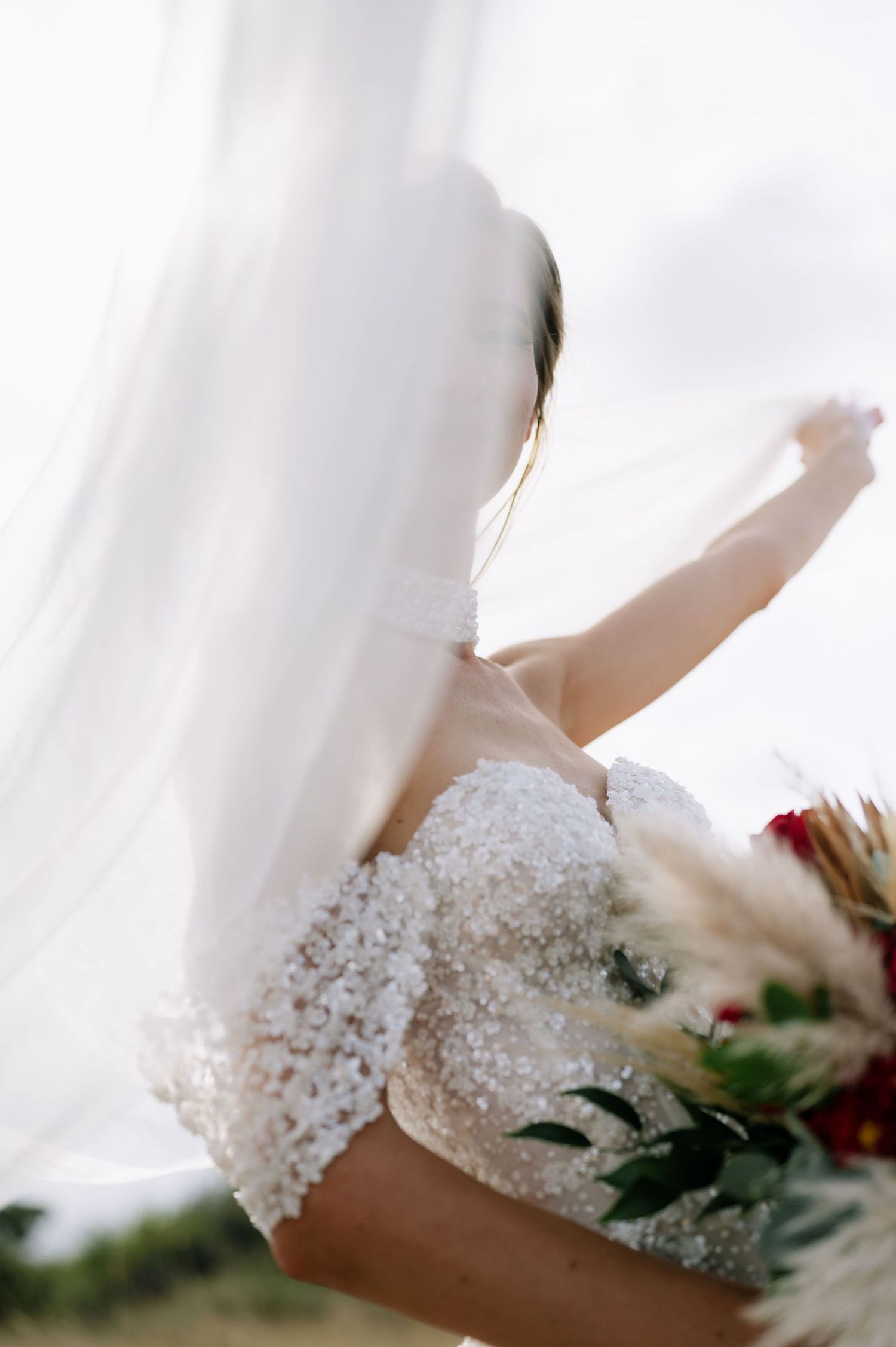 An artistic shot of a bride in a beaded gown with flowing veil against a bright background.