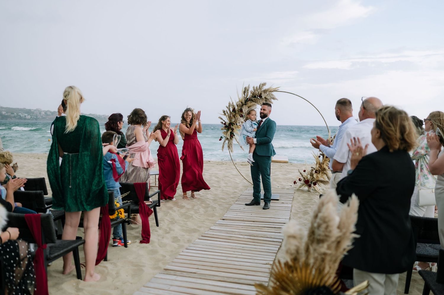 Beach wedding ceremony setup with wooden boardwalk and guests in formal attire by the ocean.