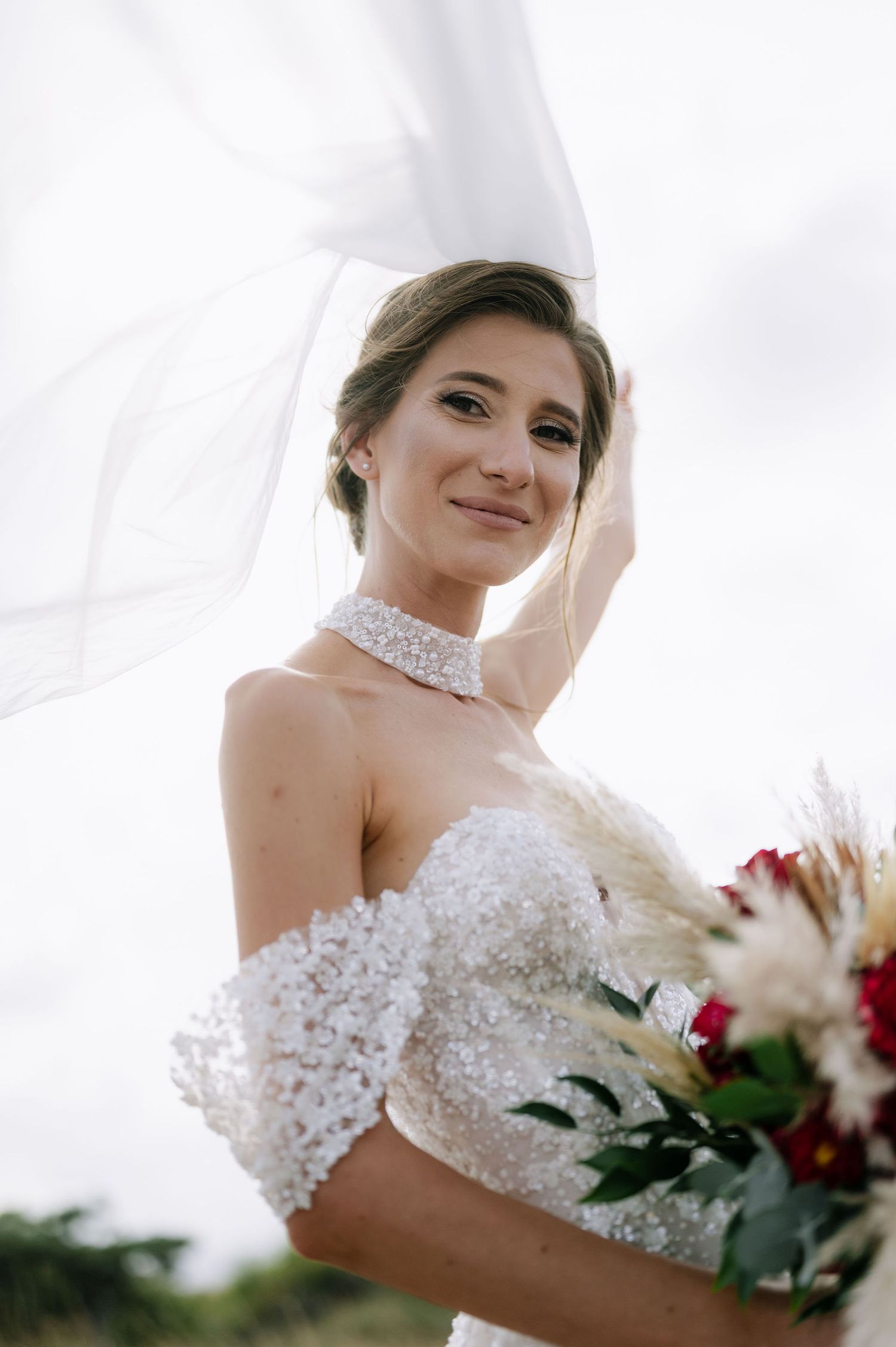 Bride in detailed lace wedding dress holds a white and red floral bouquet.