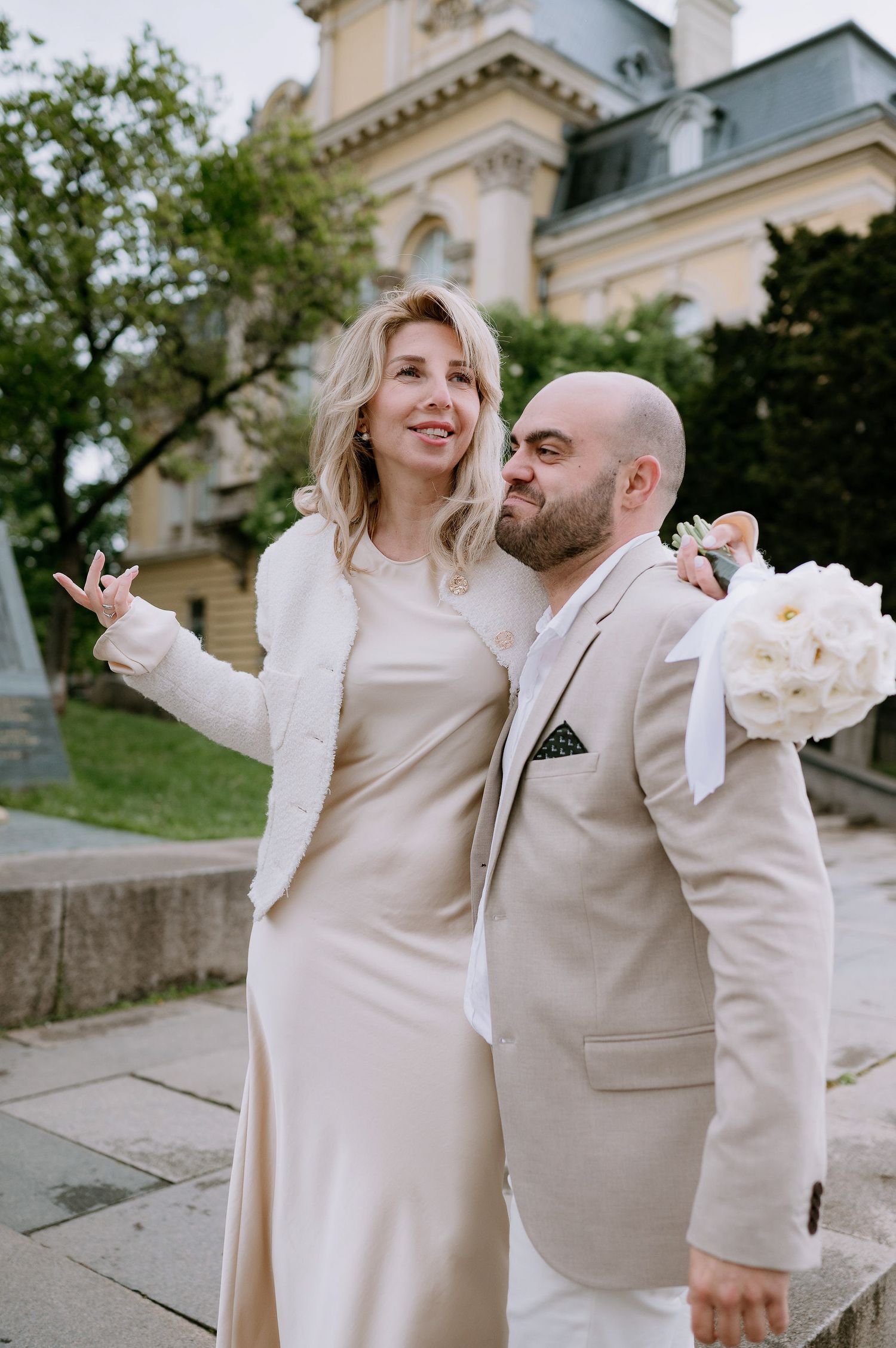 A joyful couple in cream-colored outfits share a romantic moment in front of a historic building.